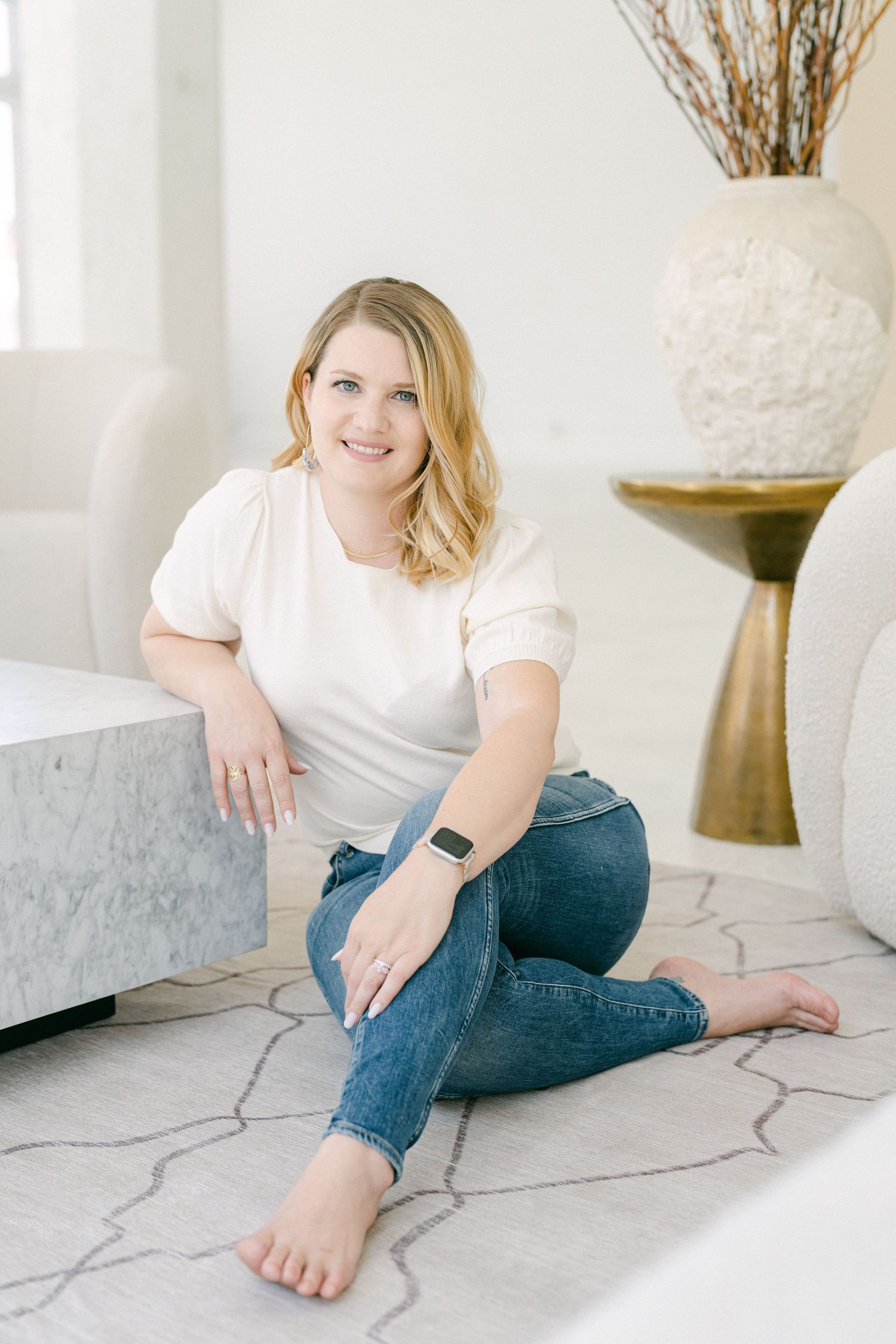 A person in white shirt and jeans sits casually on a light marble floor in a bright, minimalist setting.