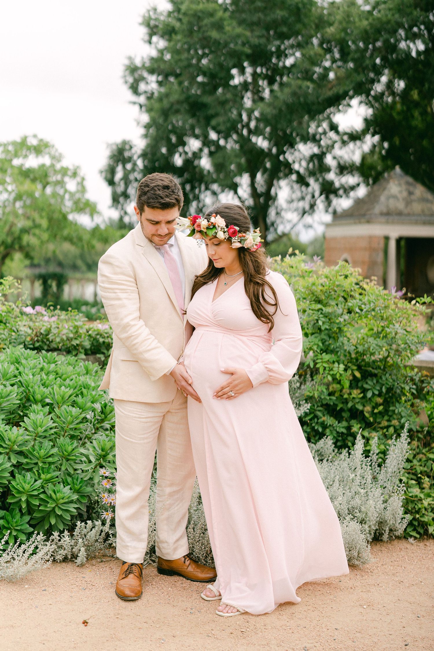 A couple in coordinating pink and cream outfits share tender moments in a garden setting with blooming flowers and greenery.