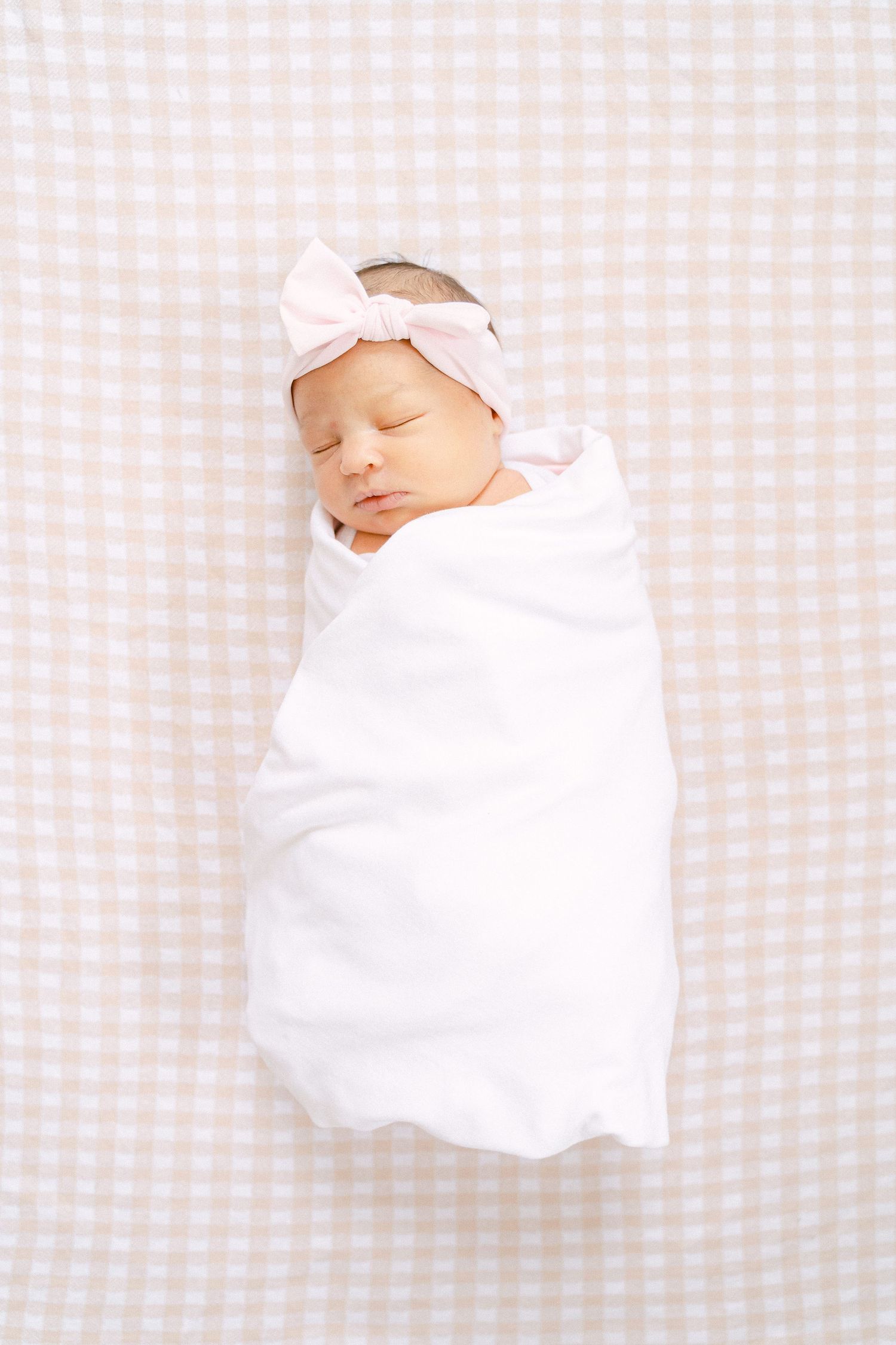 Sleeping newborn wrapped in white swaddle with pink bow headband on gingham background.