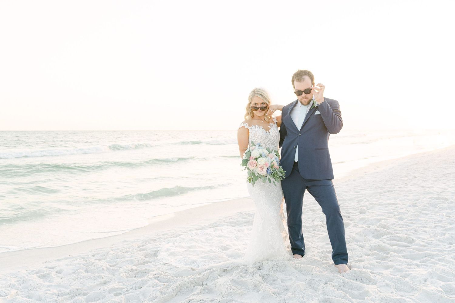 Bride and groom in sunglasses laughing together on a sunny beach, bride holding a white bouquet in Destin, FL