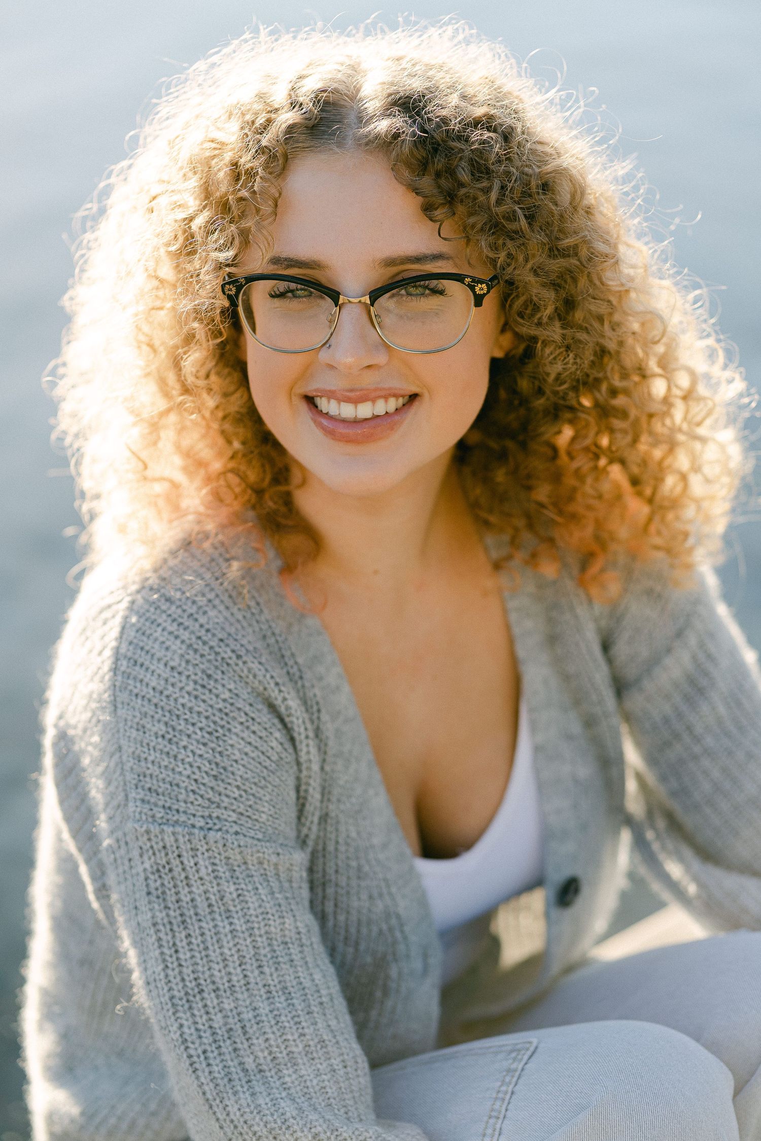 Backlit portrait of someone with curly hair wearing a gray sweater and glasses smiling in warm sunlight.