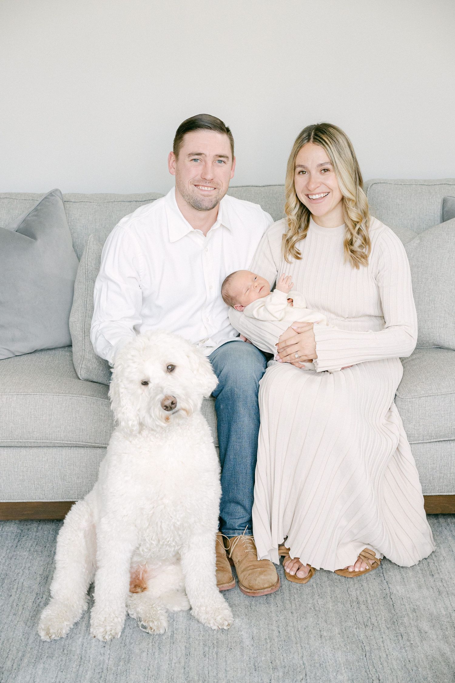 A couple sits on a gray couch with their white fluffy dog in a bright modern living room.