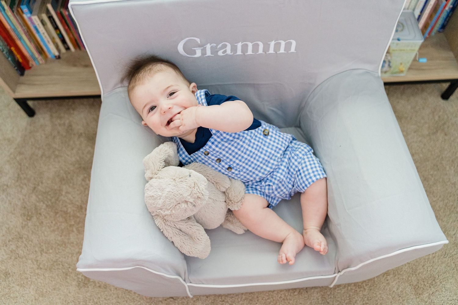 A baby rests in a light gray rocking chair next to plush toys and bookshelves.
