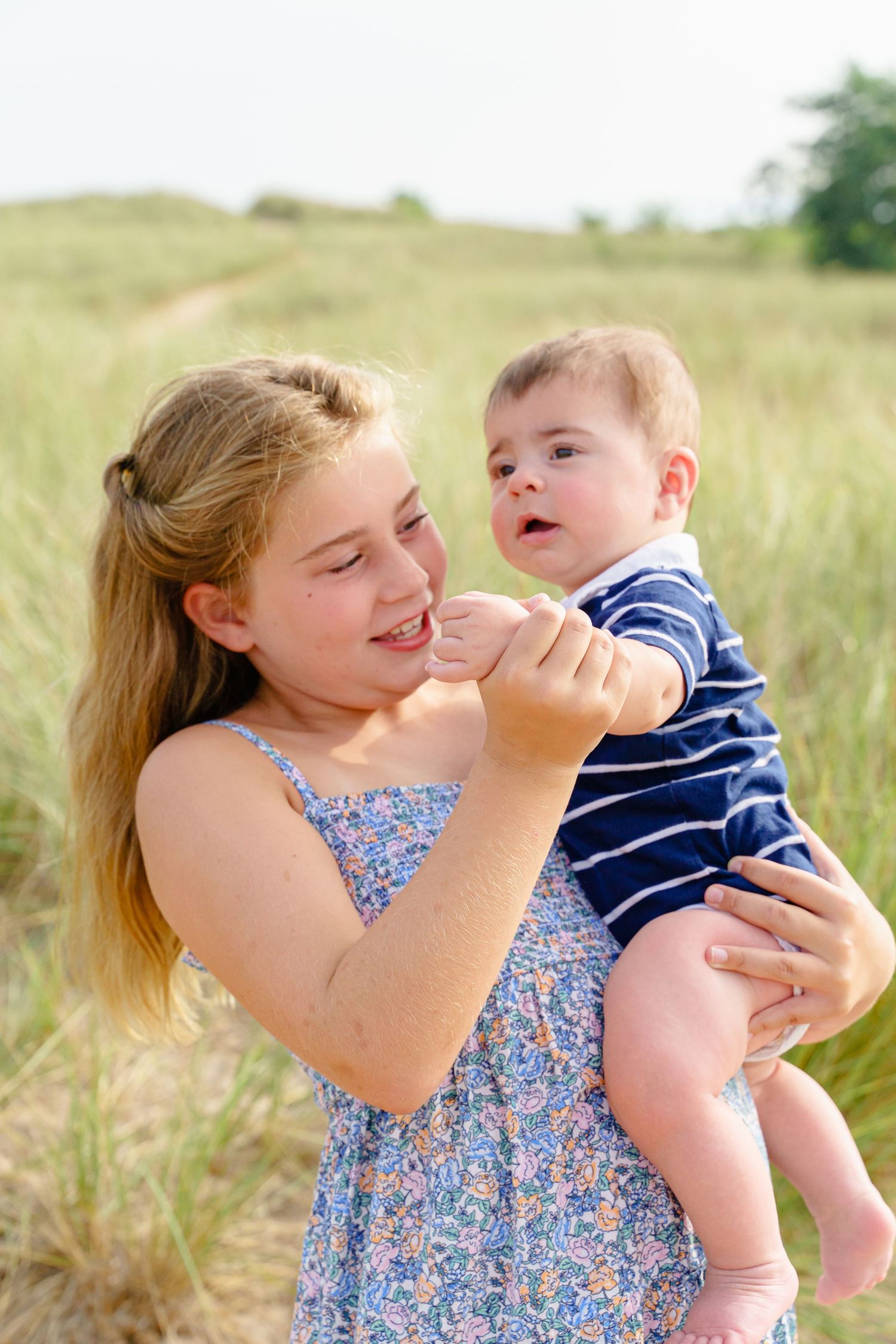 A person in a floral dress holds and plays with a baby in a navy striped outfit in a sunny outdoor field.