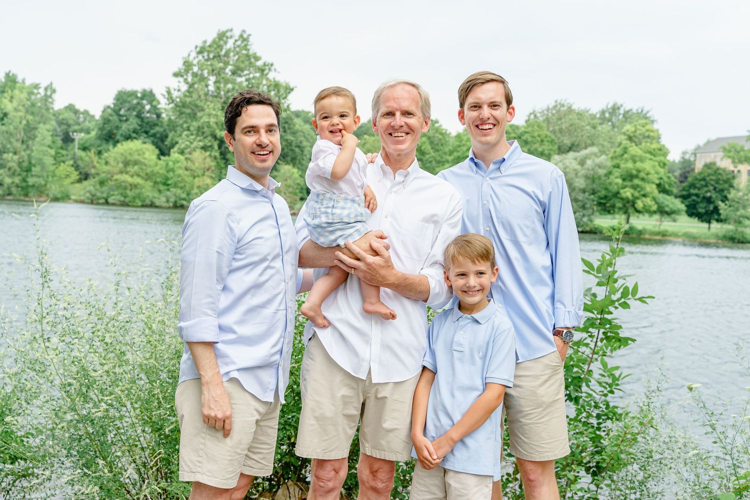 Family members gather for a casual outdoor portrait by a lakeside with trees in the background.