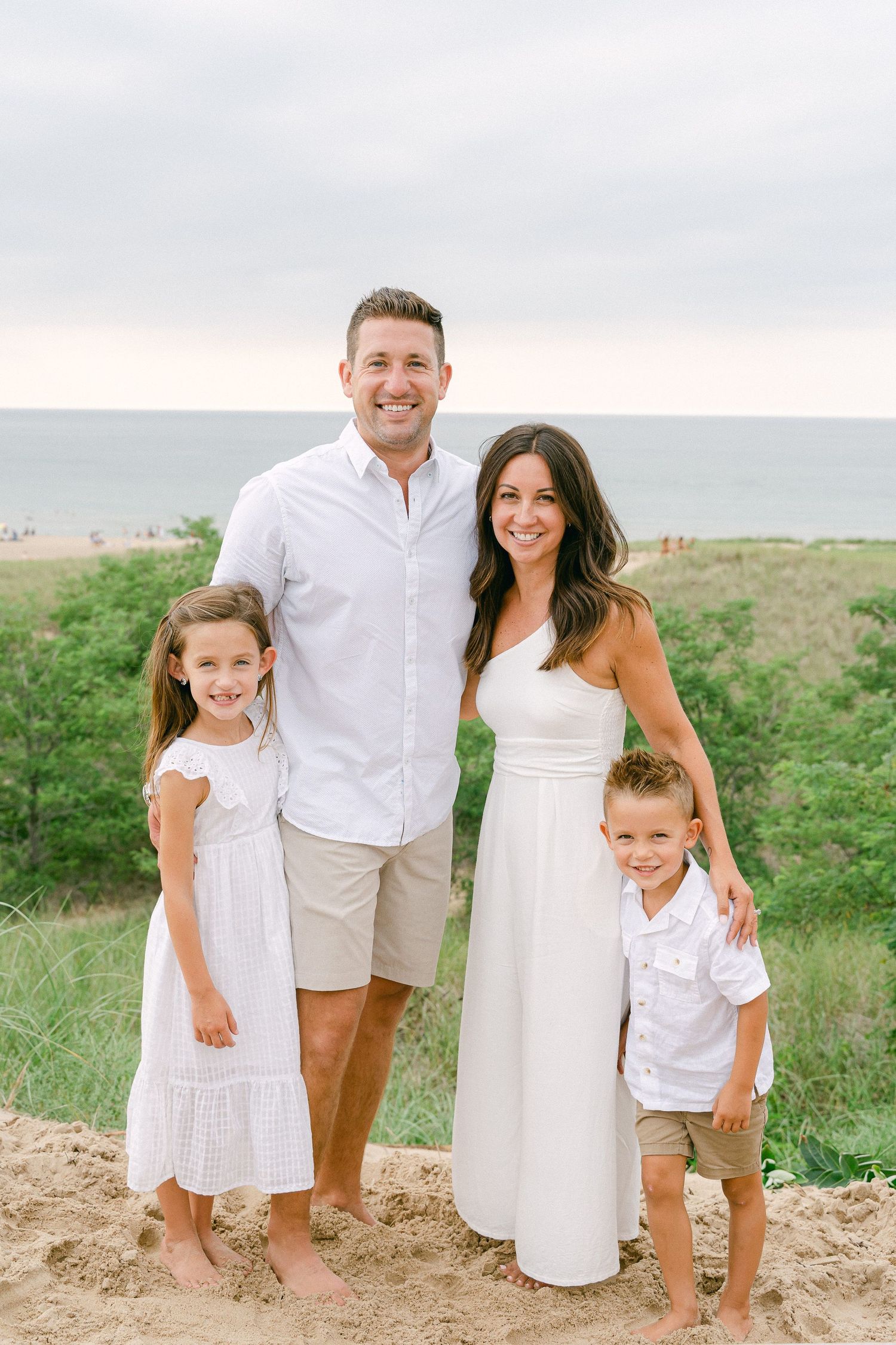 Beach family portrait with coordinated white and beige outfits against coastal scenery.