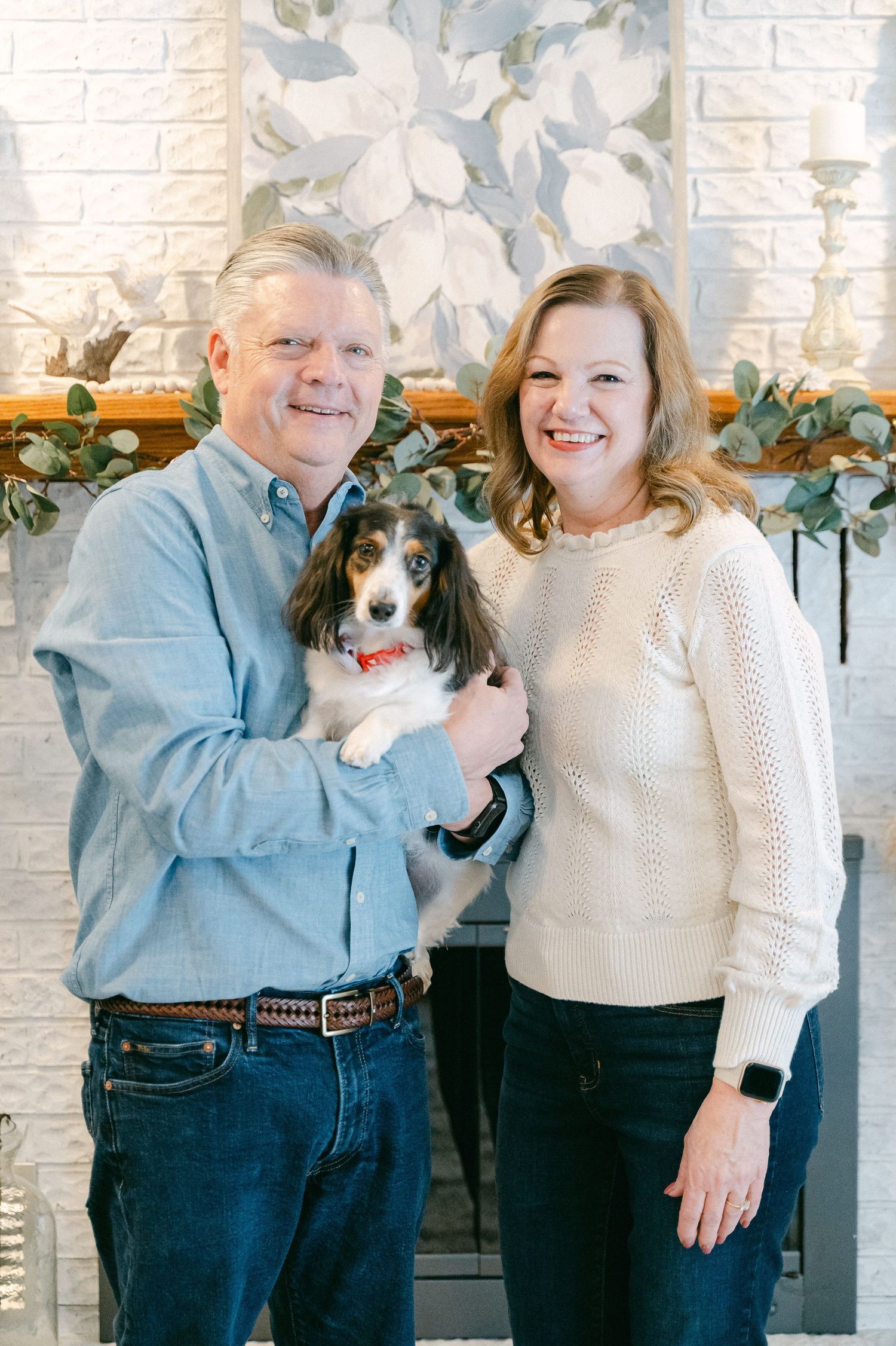 A couple poses with their small dog in front of a decorated white brick fireplace.