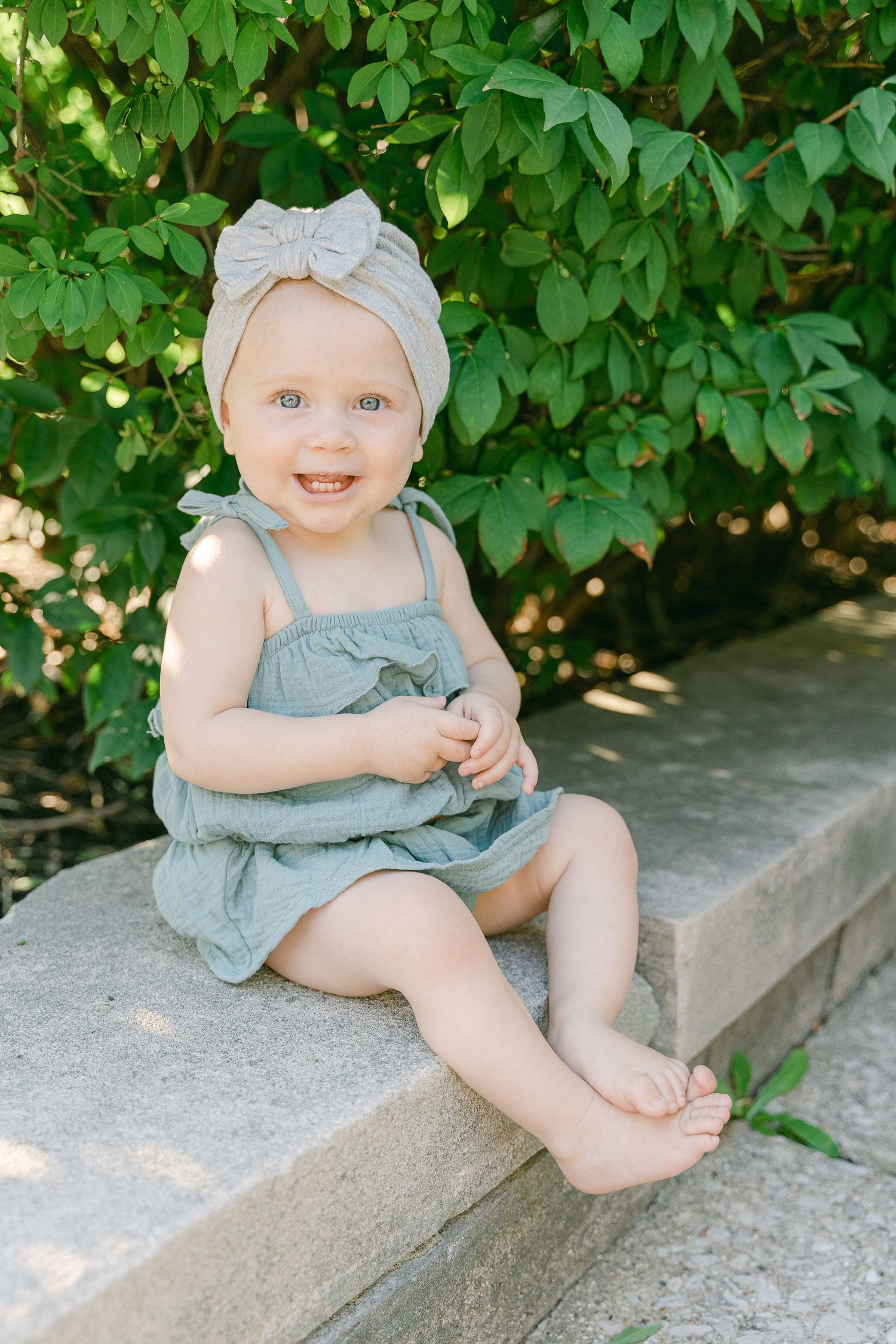 Baby in blue romper sitting on concrete ledge with green leafy background.