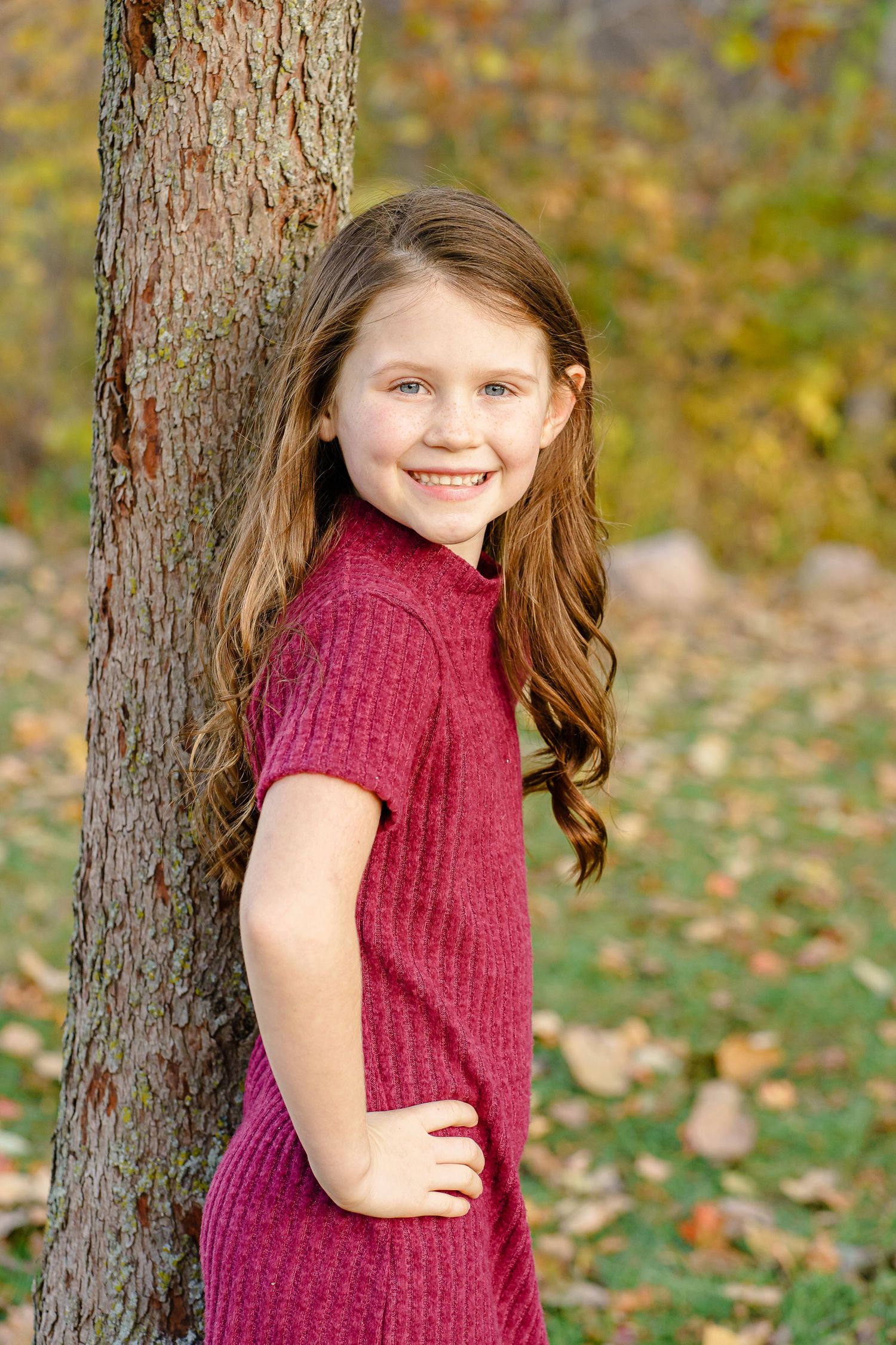 A child in a burgundy sweater poses playfully against a tree trunk with autumn leaves scattered on the ground.