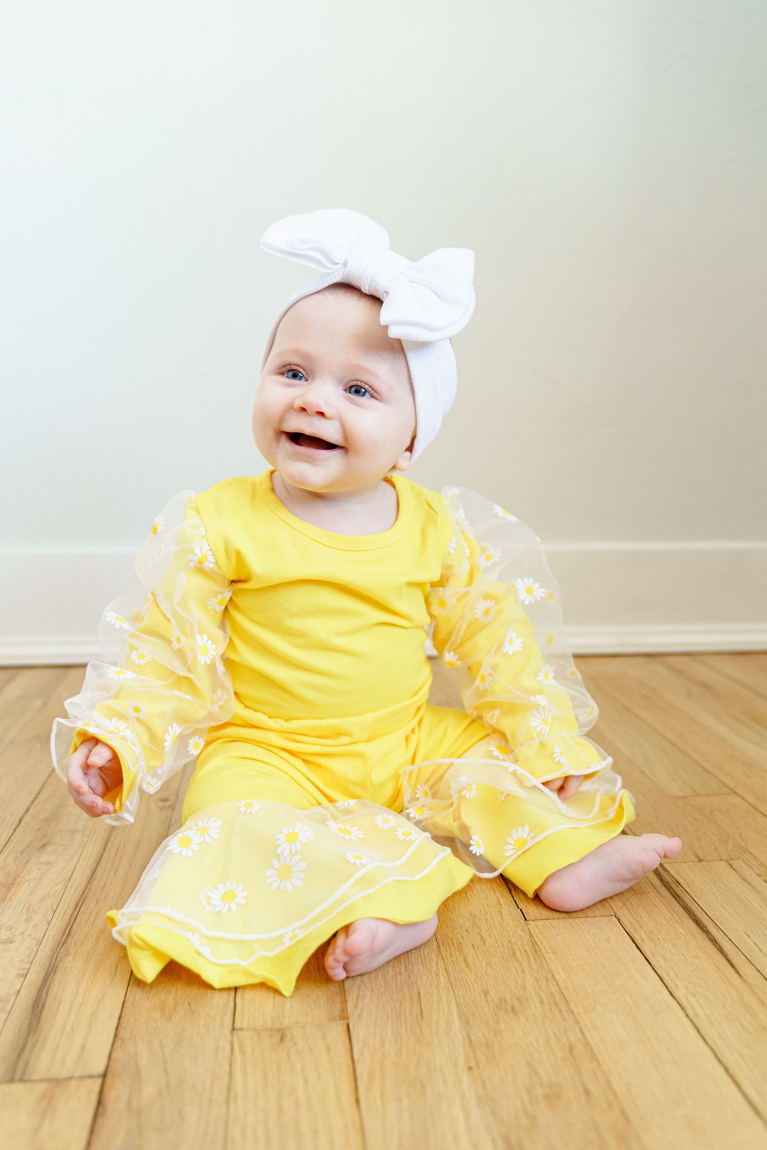 Baby in bright yellow dress with white floral overlay sits smiling on hardwood floor.