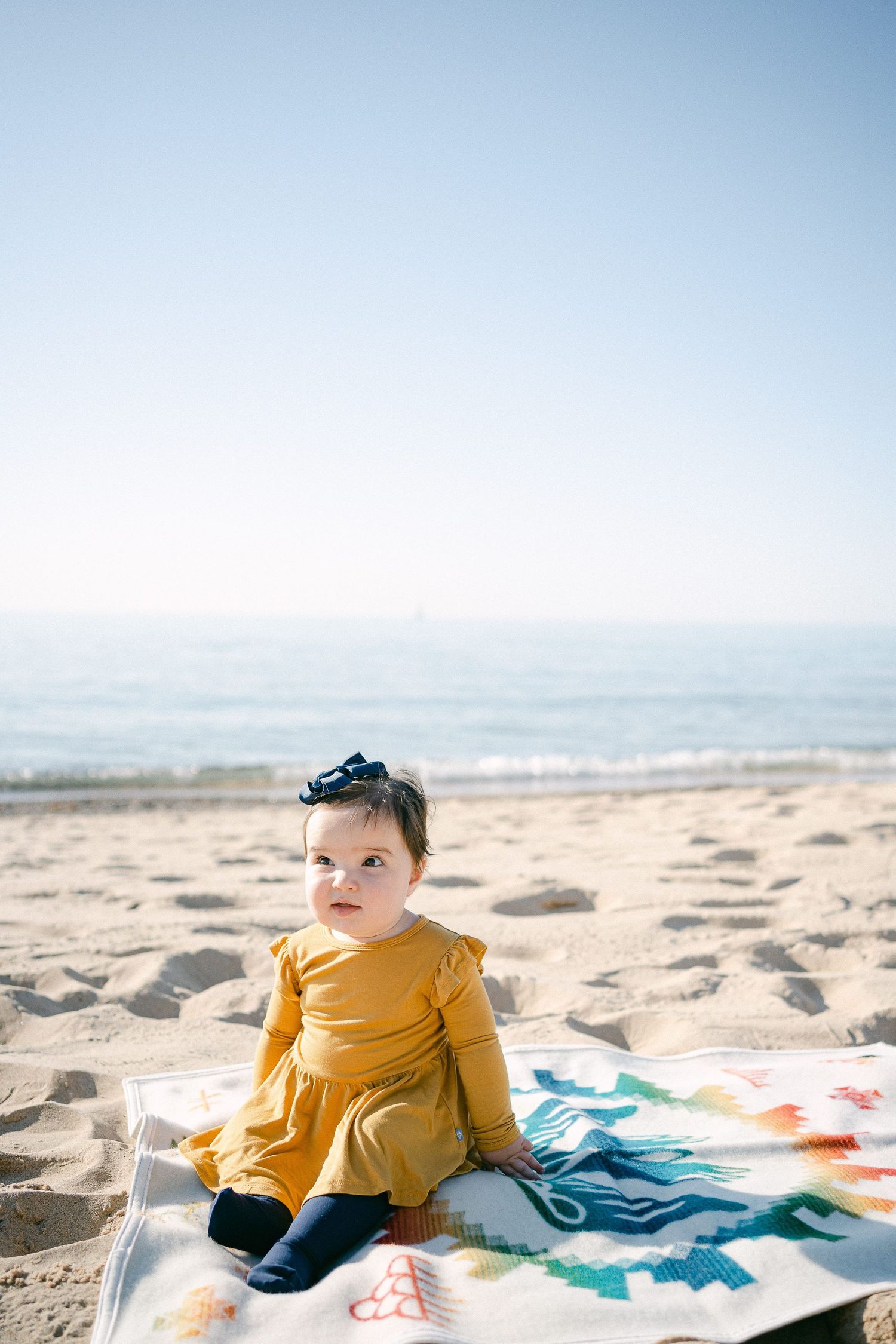 A small figure in a mustard yellow sweater sits on a colorful blanket at the beach.