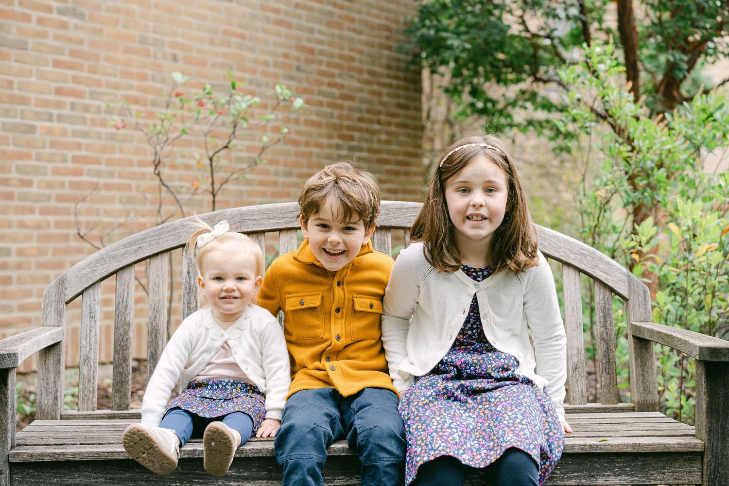 Family members sitting together on a wooden bench against a brick wall surrounded by greenery.