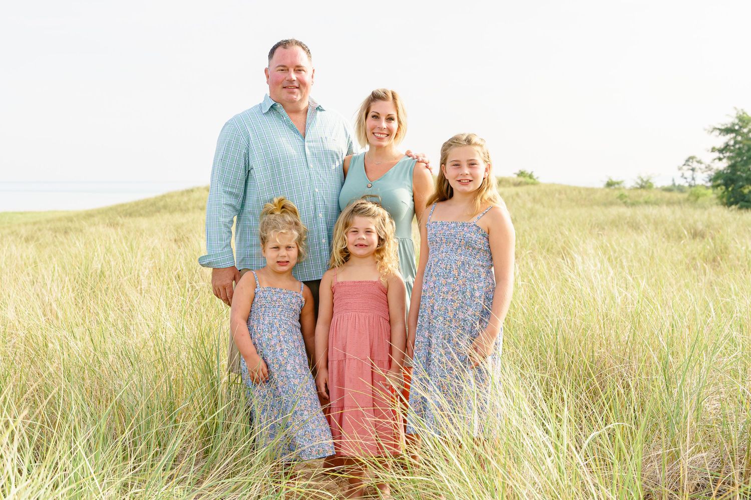A family stands together in a sun-lit grassy field wearing coordinated pastel and blue outfits.