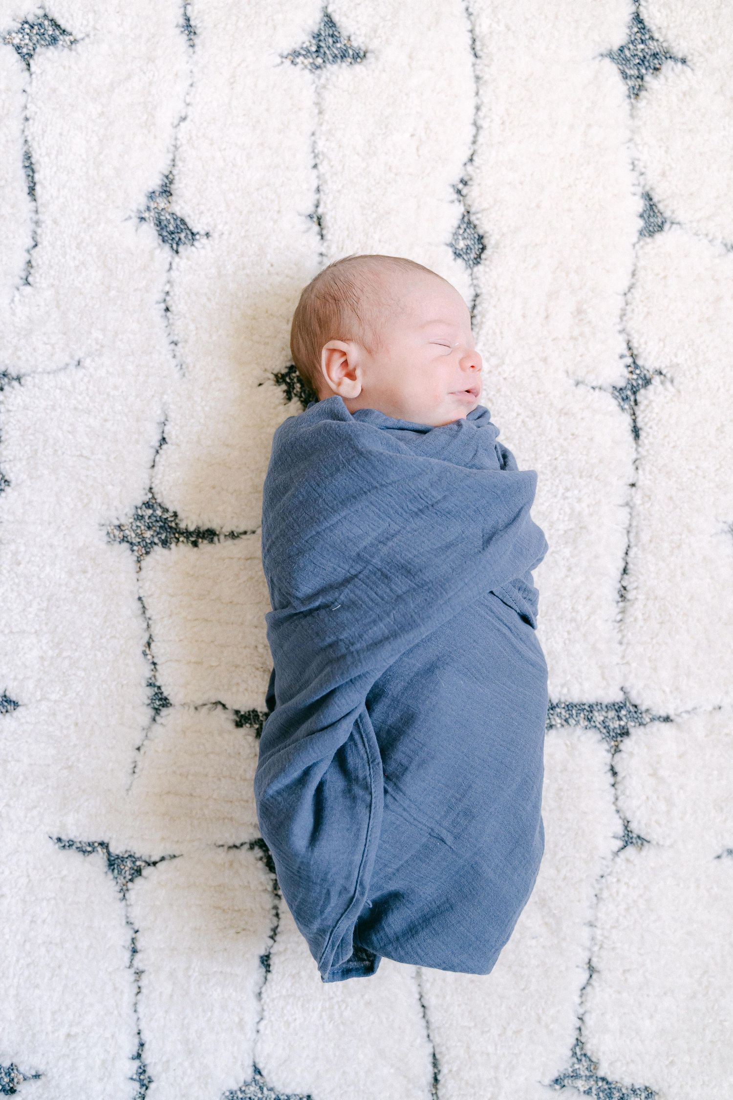 A sleeping newborn baby wrapped in a blue swaddle blanket lies on a white textured surface with butterfly pattern.
