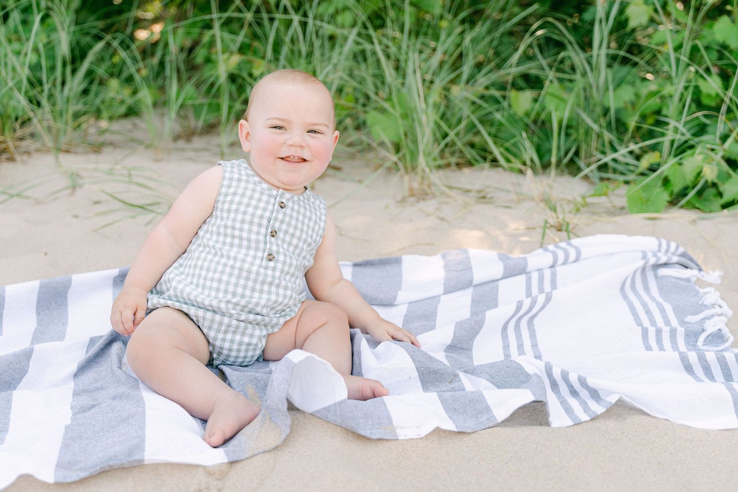 Baby in blue and white gingham romper sits on striped blanket in outdoor garden setting.