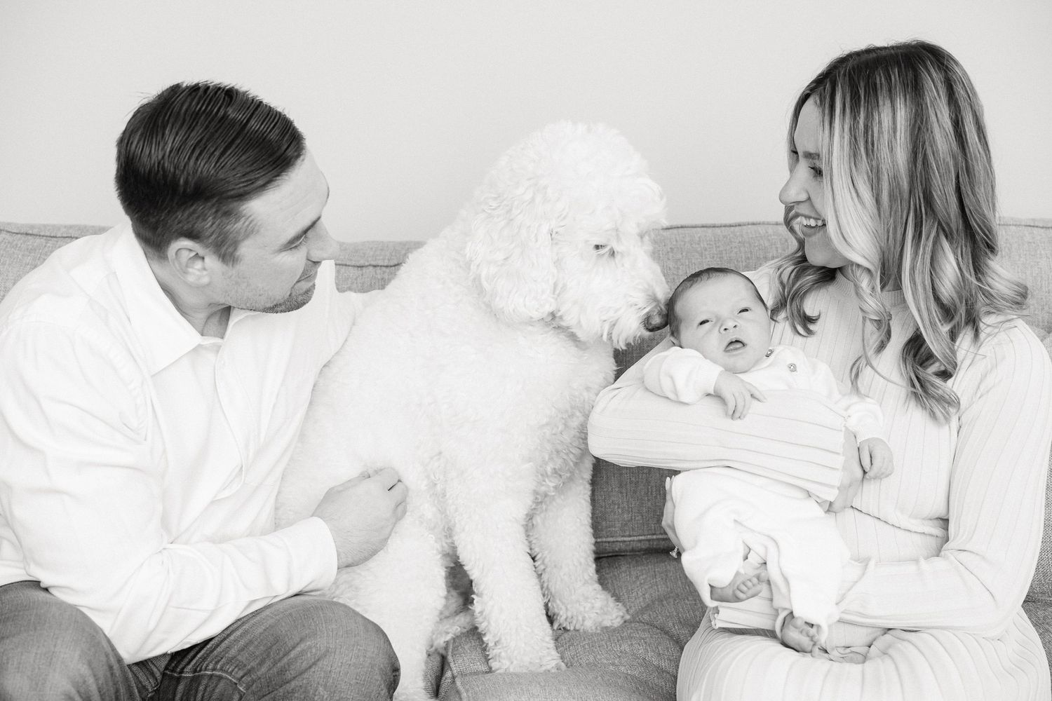 A family sits on a couch with their white fluffy Goldendoodle dog and newborn baby in a black and white photograph.