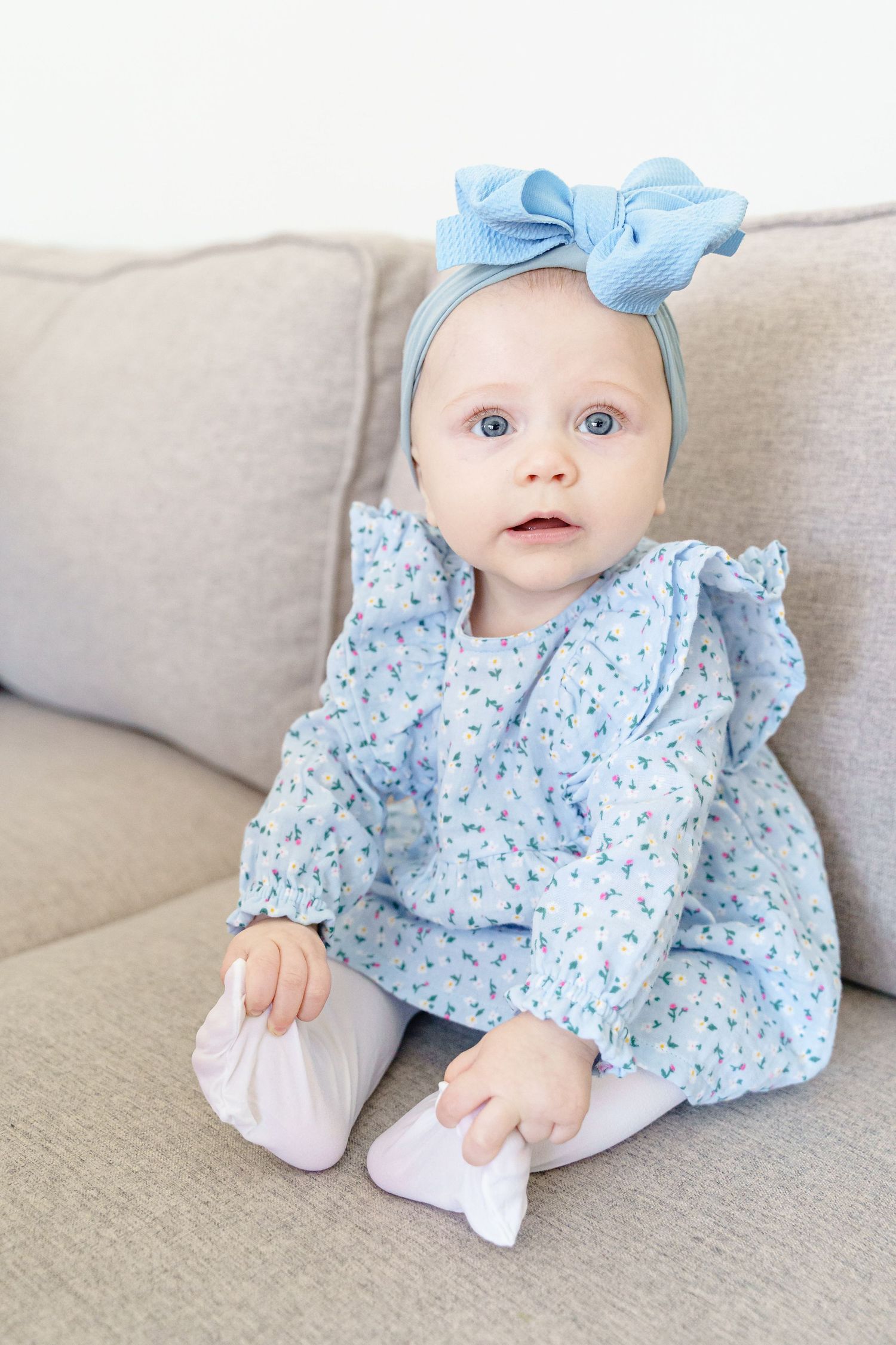 Baby wearing light blue floral dress and bow headband sitting on beige couch.