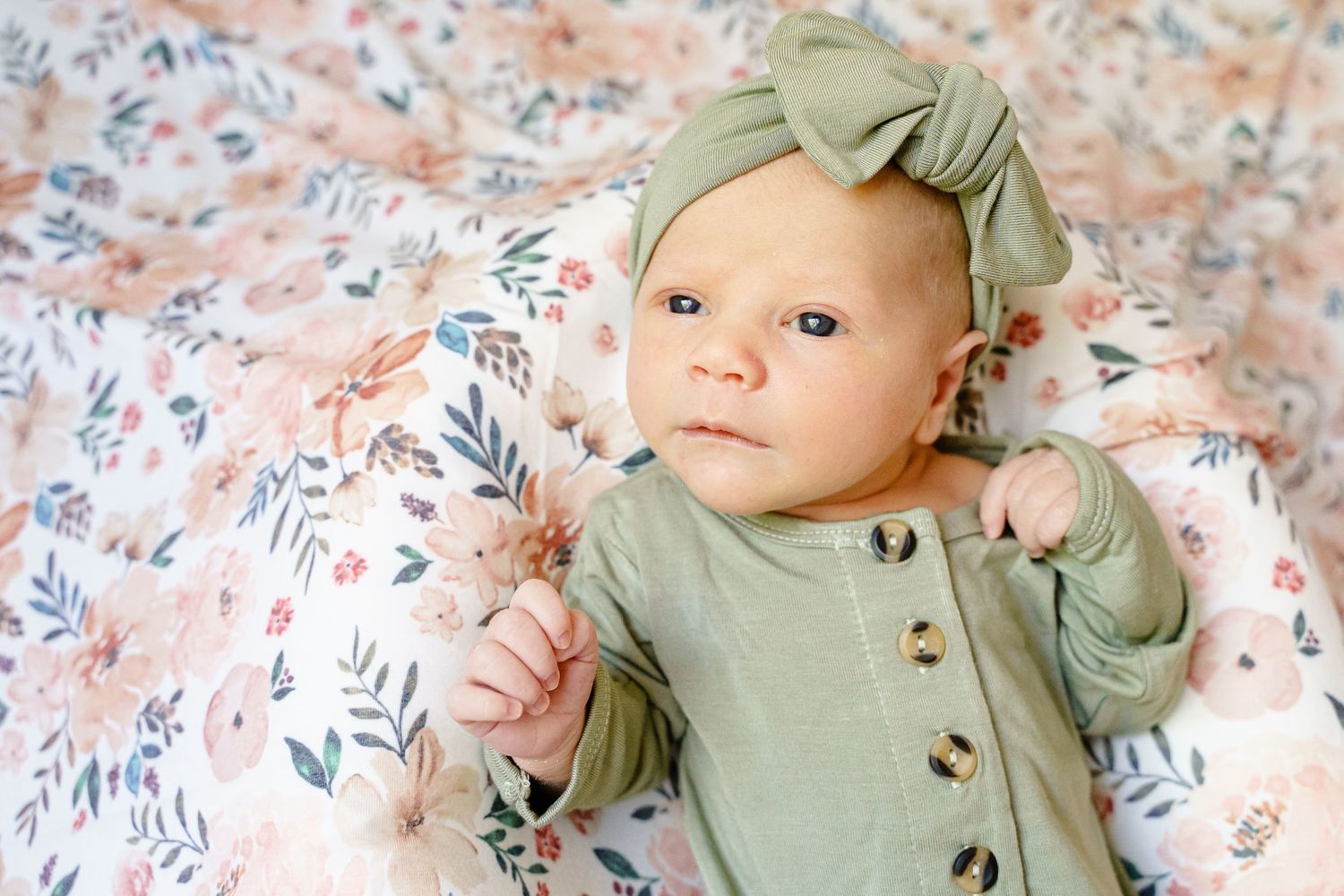 Baby in sage green outfit with matching bow lying on floral print fabric.