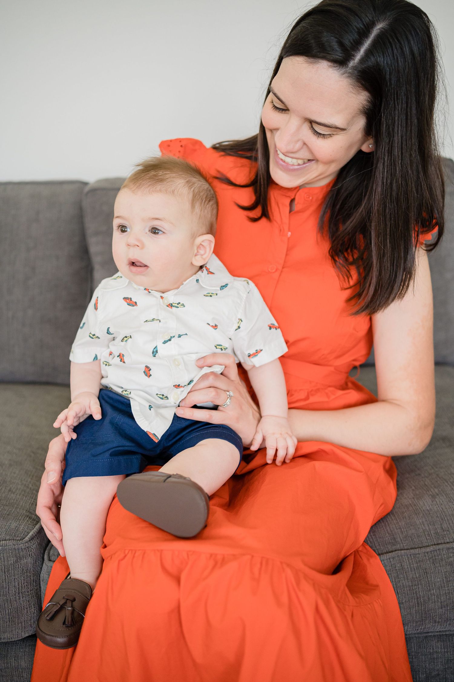 Mother in bright orange dress sits on grey couch with baby wearing white onesie and navy pants.