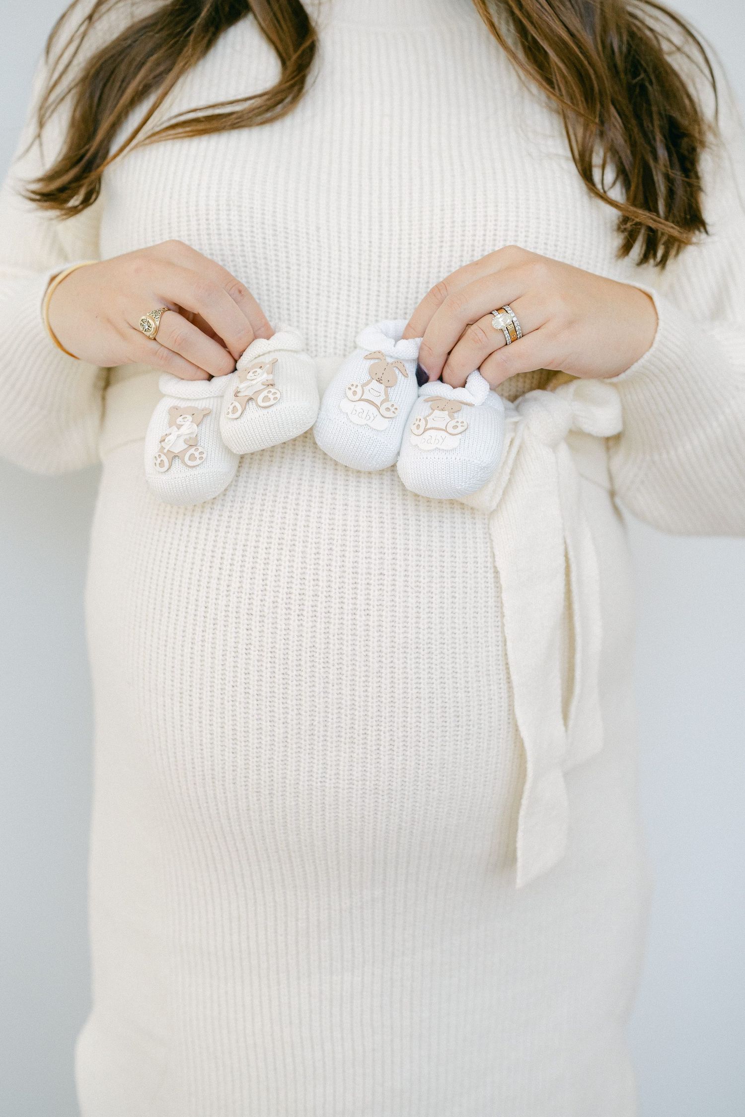 Pregnant woman in white sweater holds tiny baby shoes against her belly.