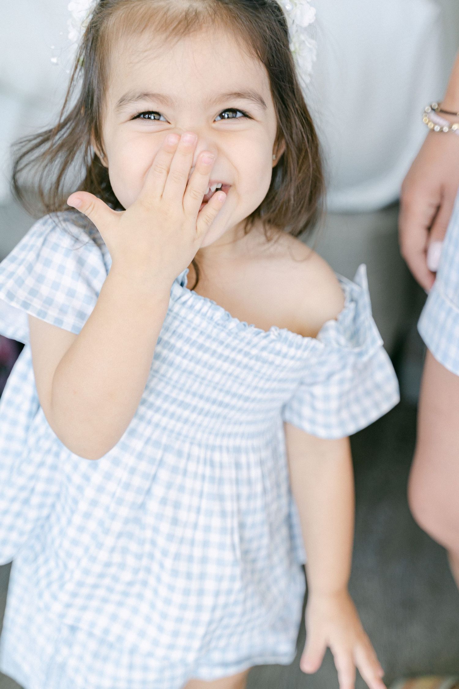 Adorable toddler girl in blue gingham off-shoulder dress laughing and covering her mouth with her hand in Miami, FL