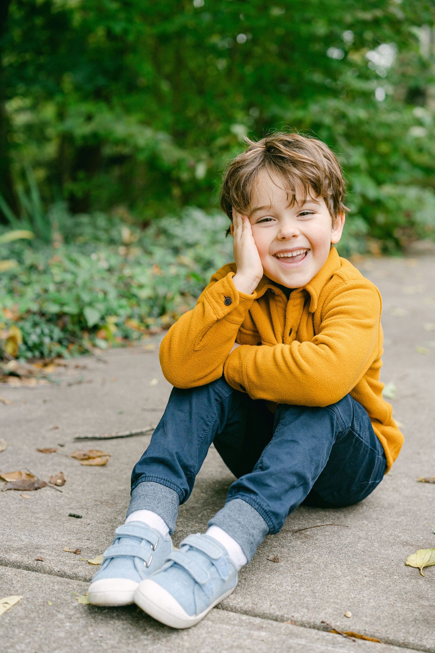 Child in yellow sweater and jeans lying playfully on a sidewalk with greenery in the background.