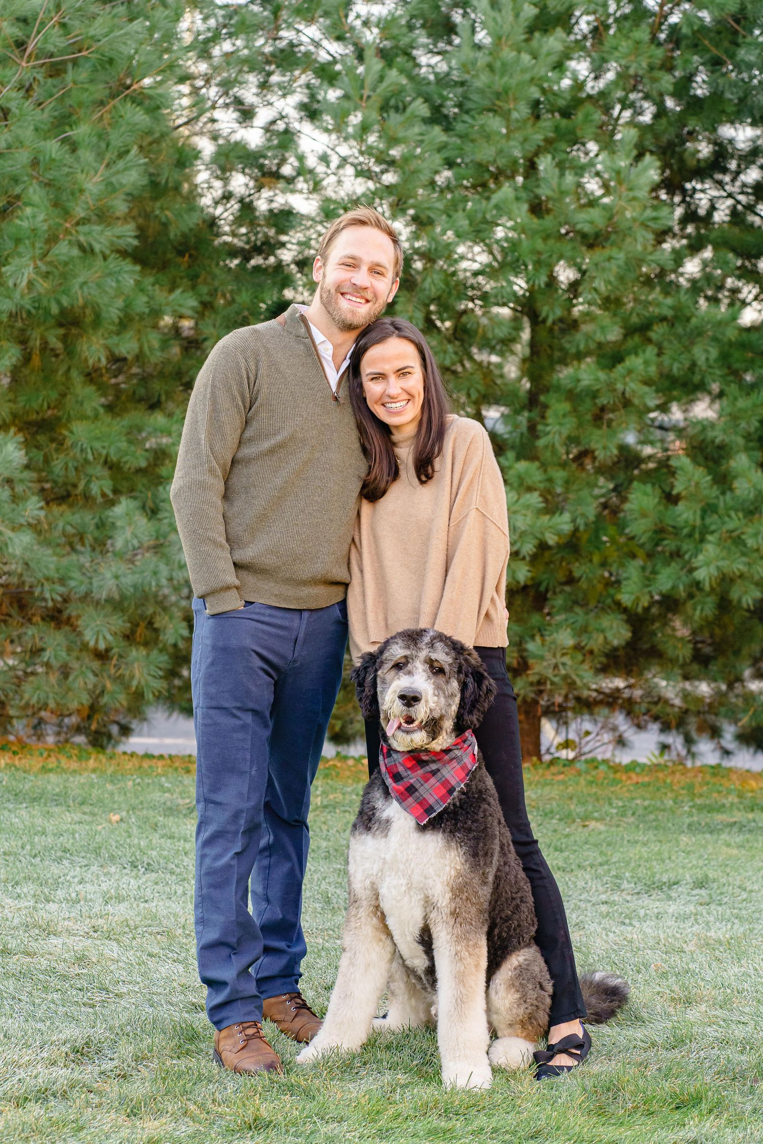A couple poses outdoors with their large mixed-breed dog wearing a red bandana among leafy green trees.