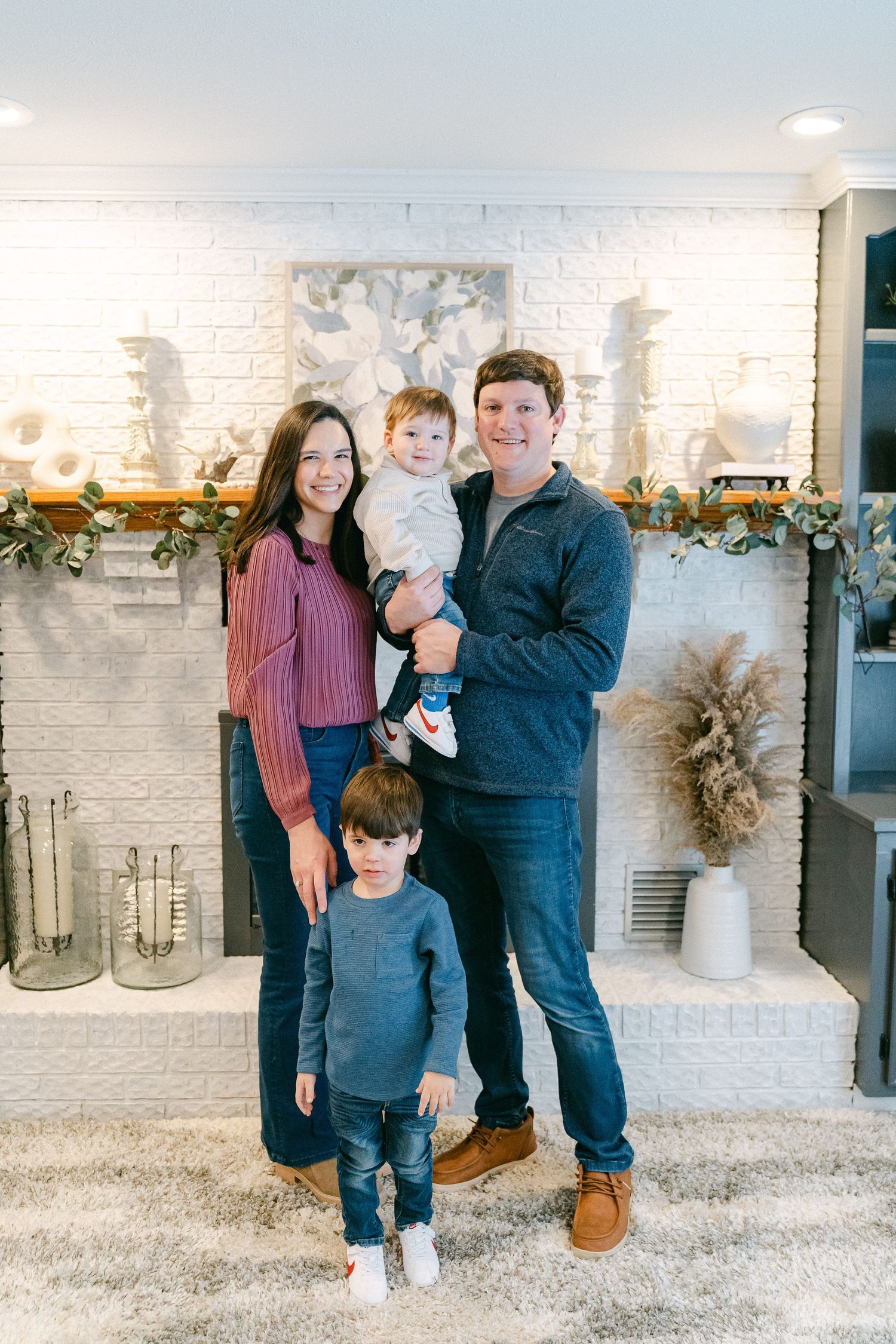 Family gathering in casual attire stands together by white brick wall with decorative garland and vase.