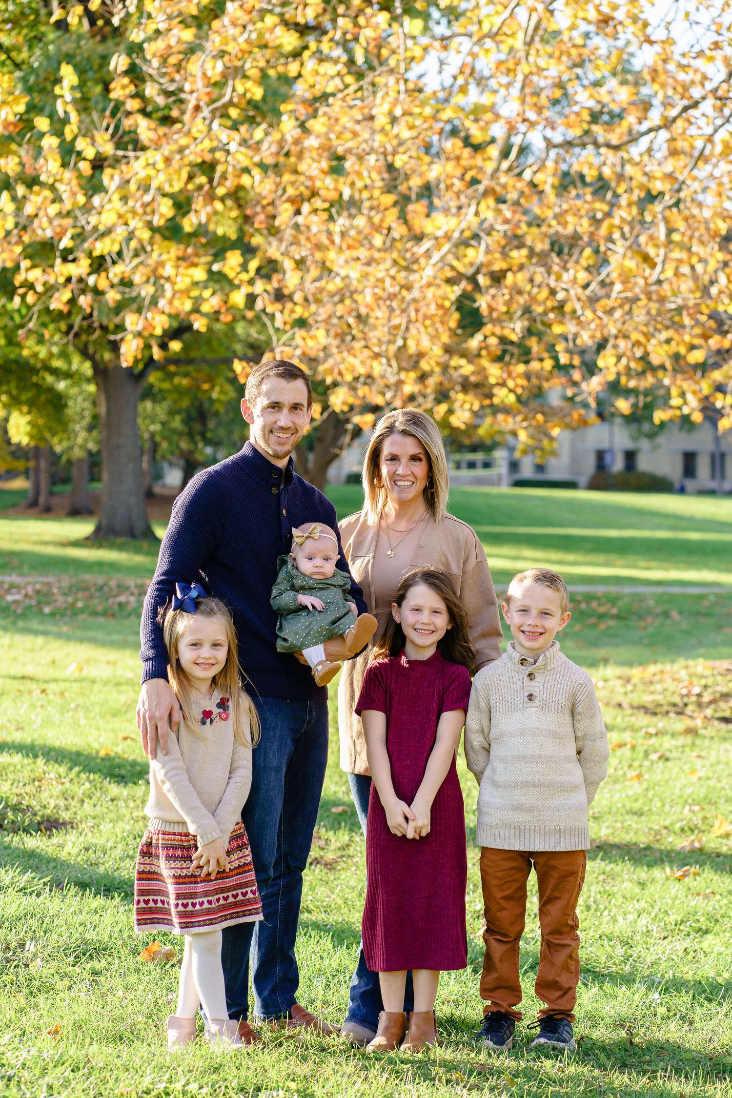 A family of five poses together outdoors on an autumn day with golden leaves in the background.