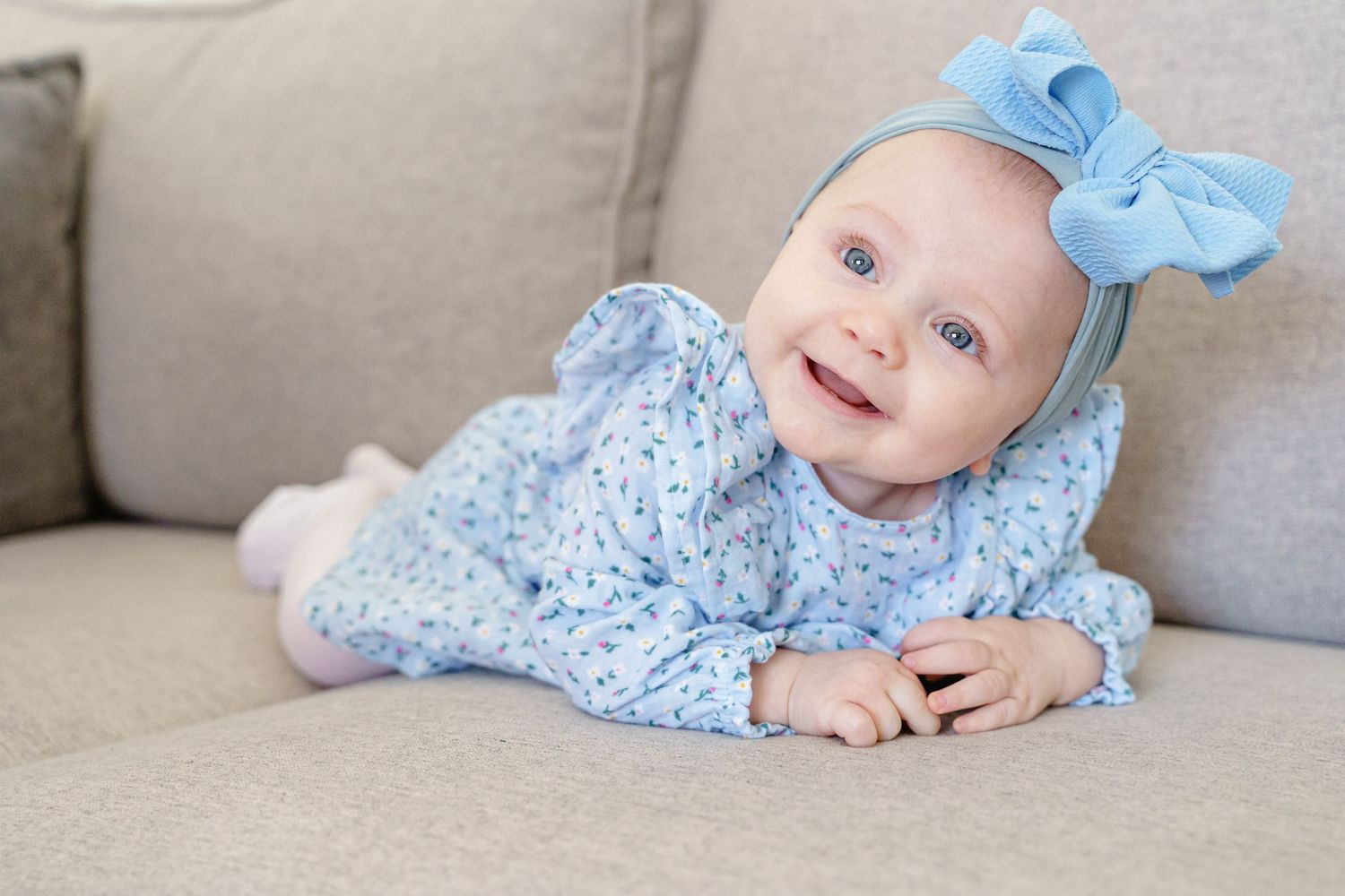 Baby in light blue lace dress and matching bow headband lying on gray couch smiling at camera.