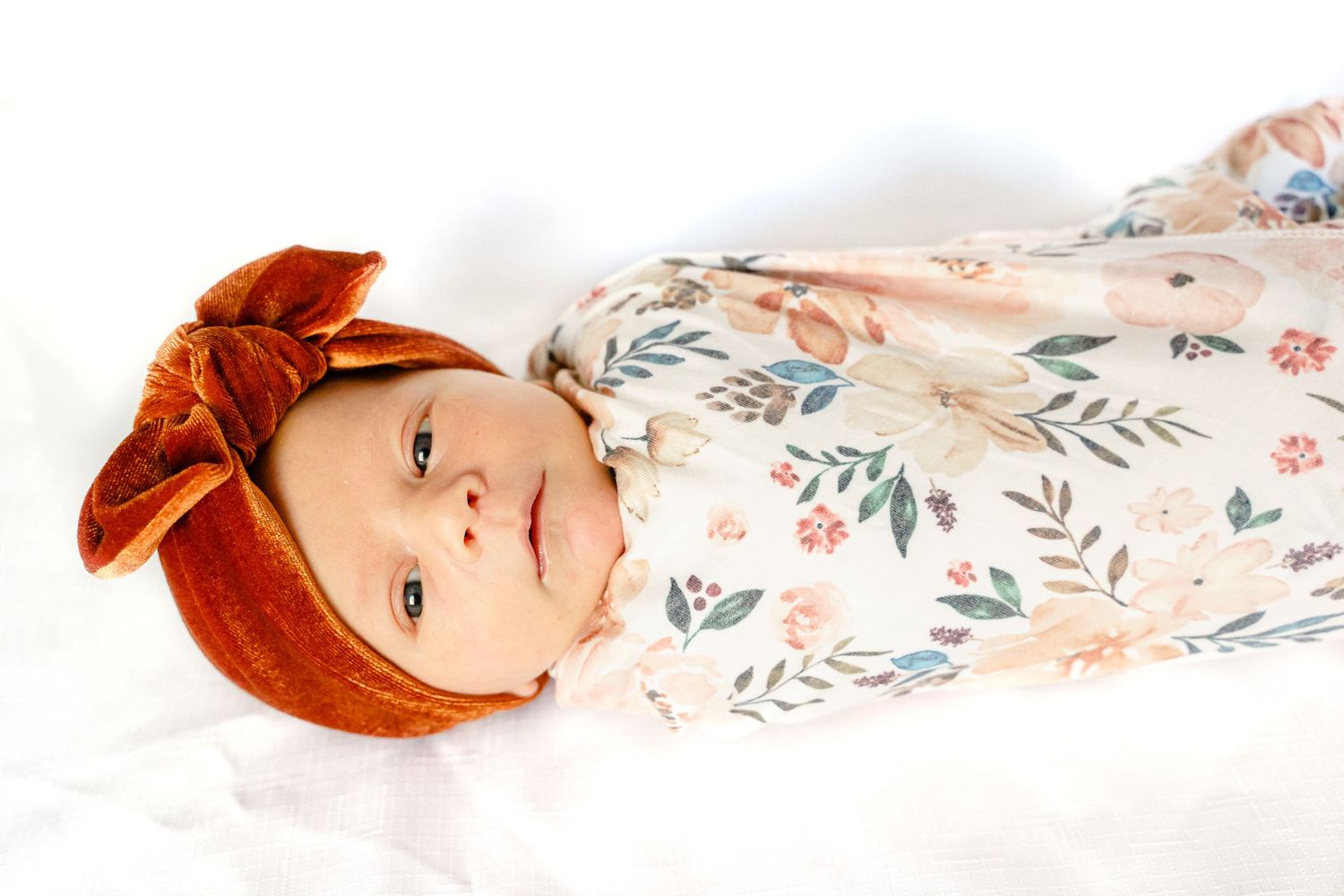 Baby wearing a rust-colored velvet bow headband lies on floral print blanket.