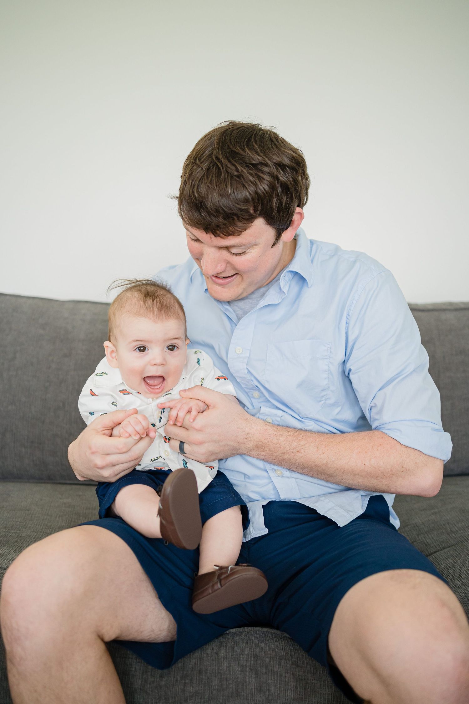 A parent in a blue shirt plays and interacts with a baby wearing a white onesie on a gray couch.