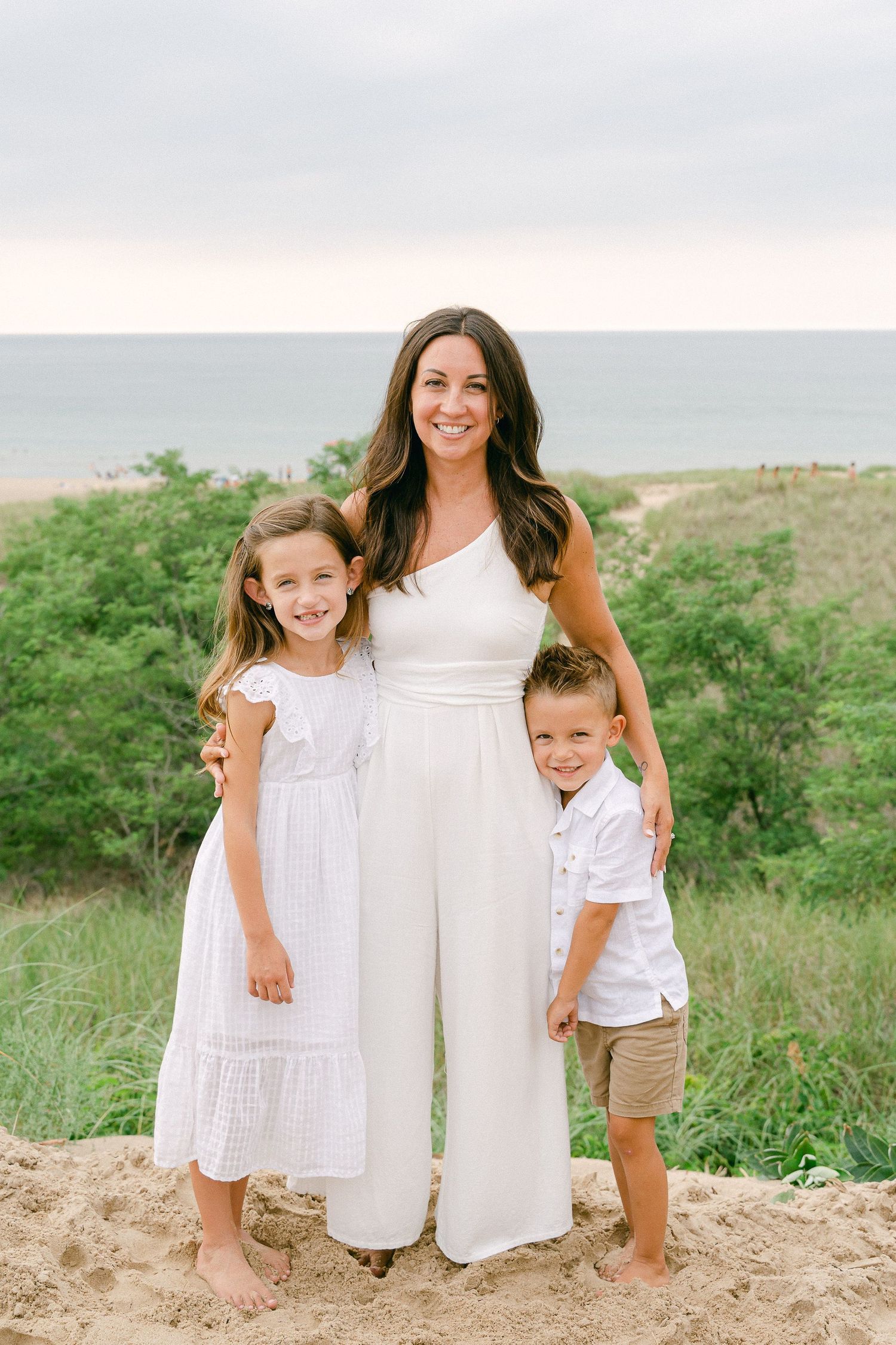 Three people in coordinating white outfits pose together on a sandy hill overlooking the ocean and greenery.