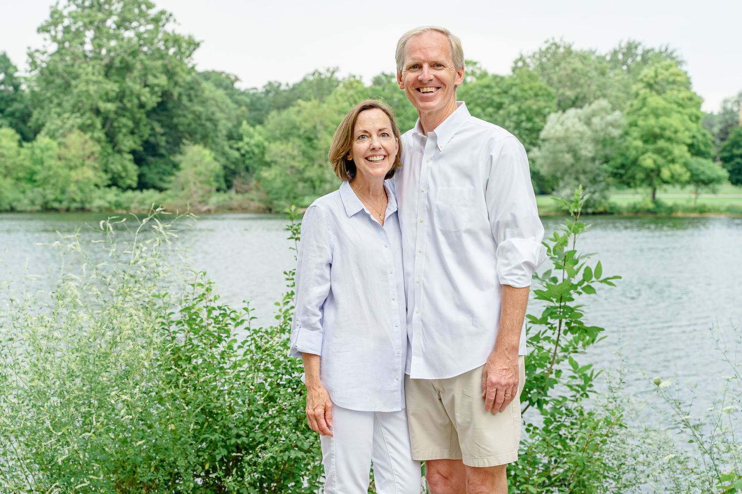 Family members in light summer clothing gather for a portrait by a scenic lake with trees in the background.