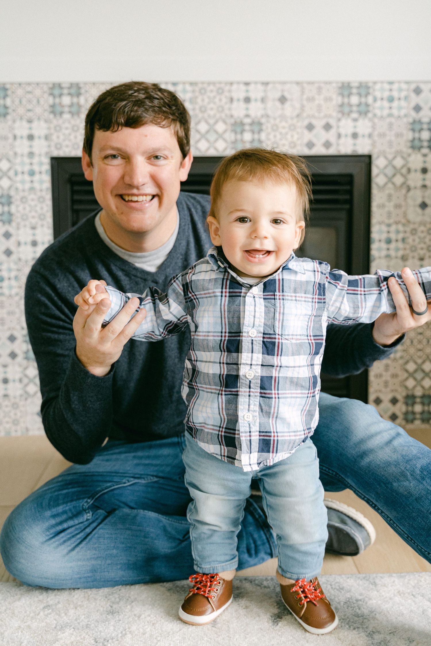 A parent and toddler sit together on a carpet, both smiling while wearing casual clothing near a decorative wall.