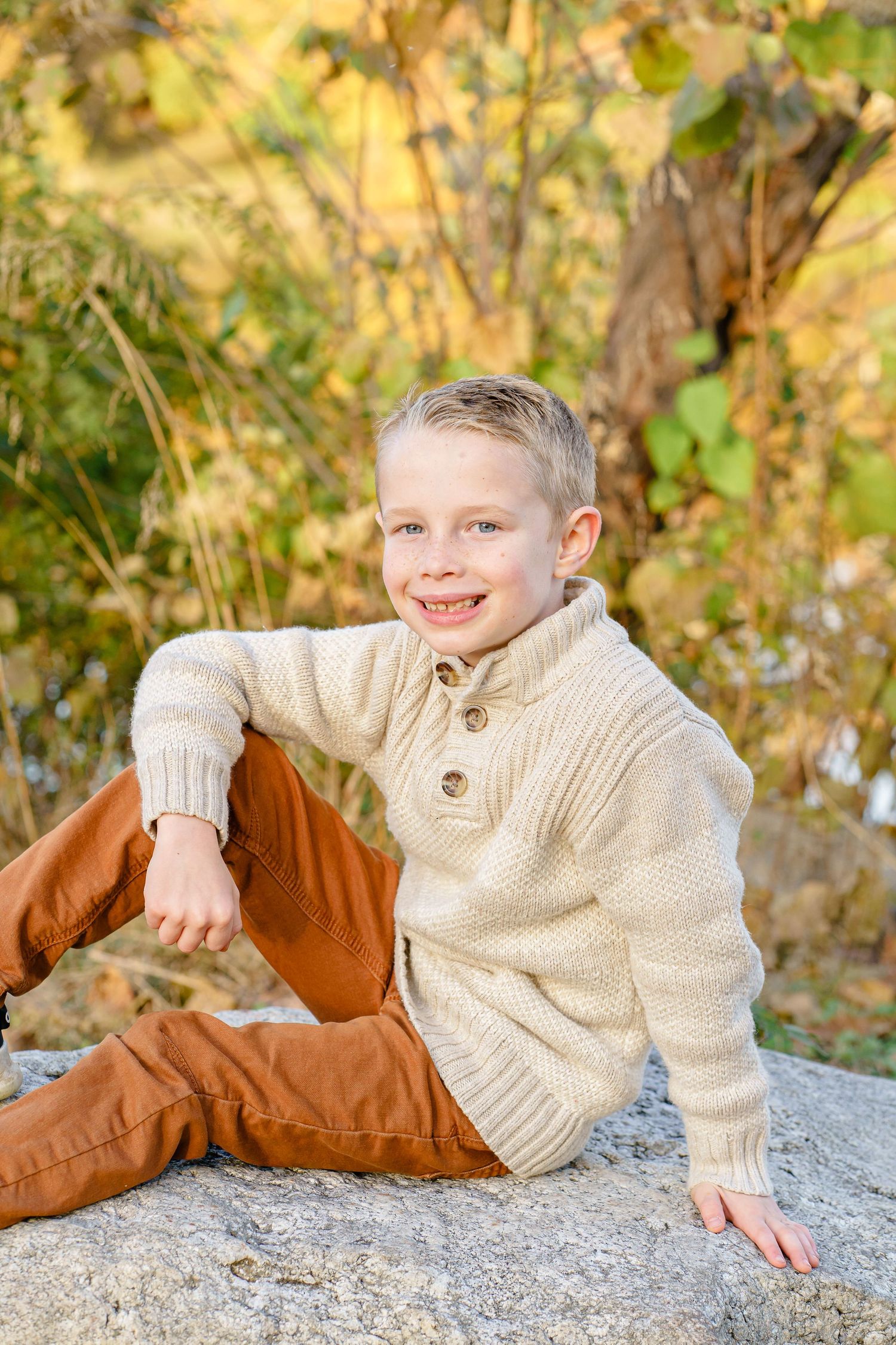 A child in a cream sweater sits casually on a rock outdoors during fall.