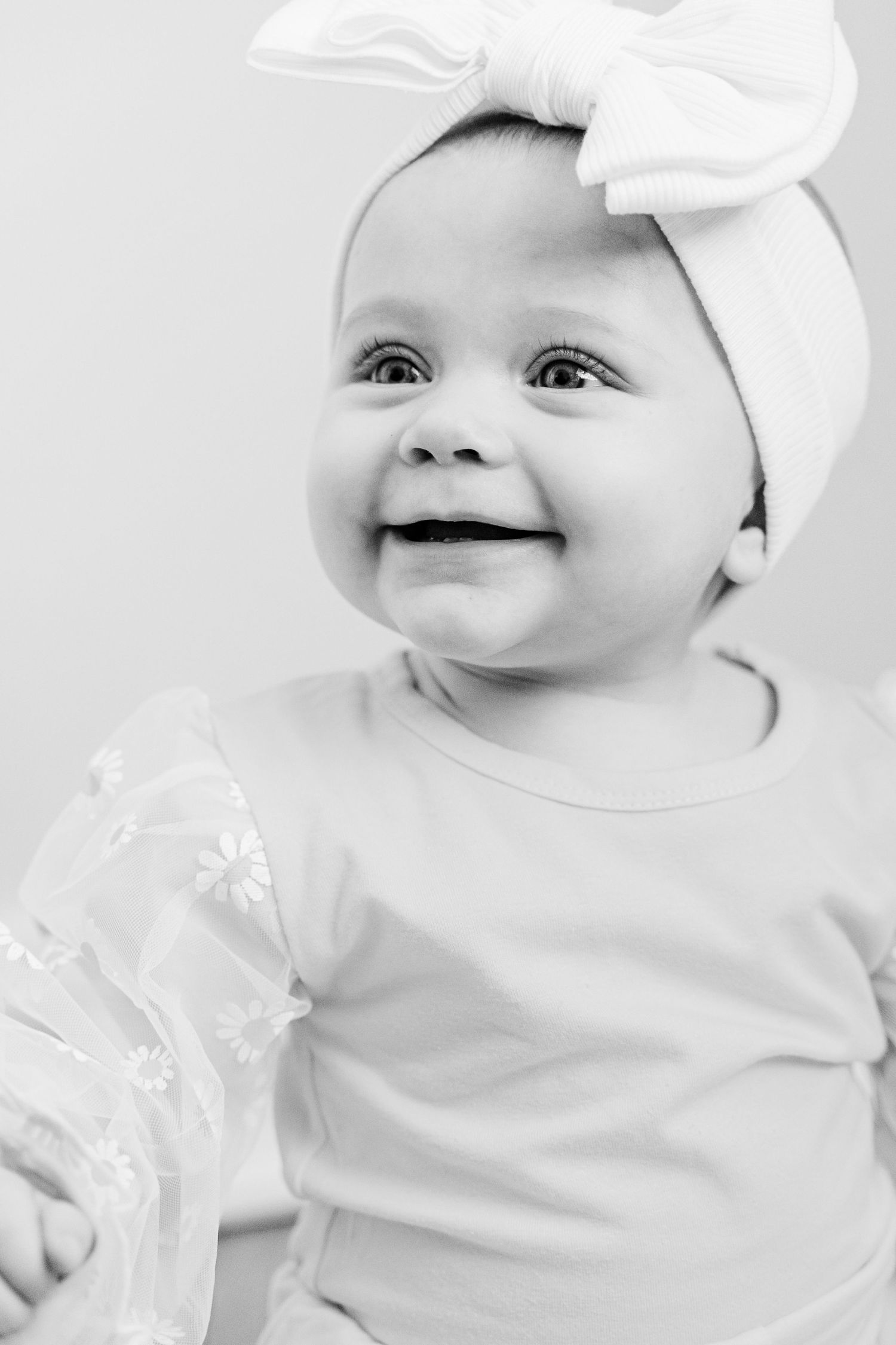 Black and white portrait of a smiling baby wearing a white bow headband and light colored outfit.