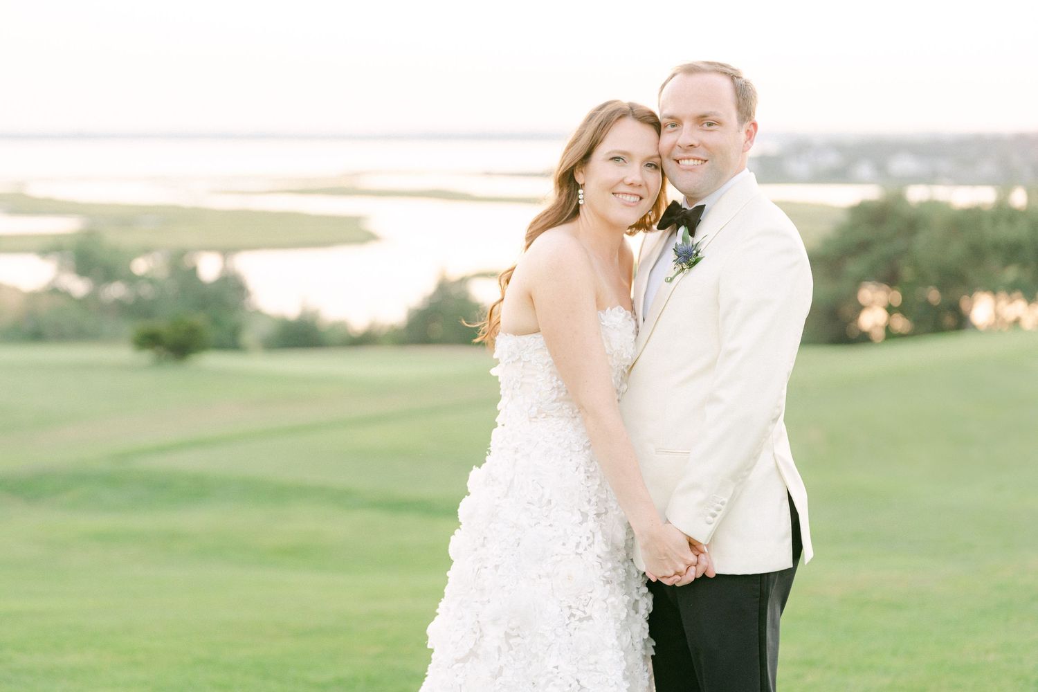 Bride and groom smiling together on a golf course at sunset, she in a ruffled gown, he in a white tuxedo jacket in Cape Cod