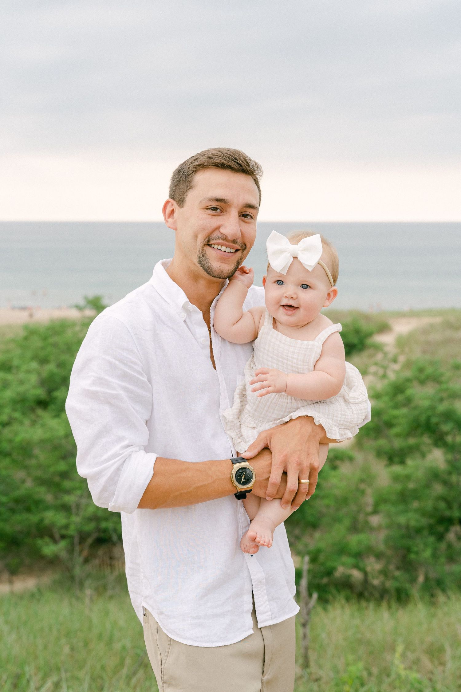 A parent in a white shirt holds and plays with a baby against a coastal landscape backdrop.