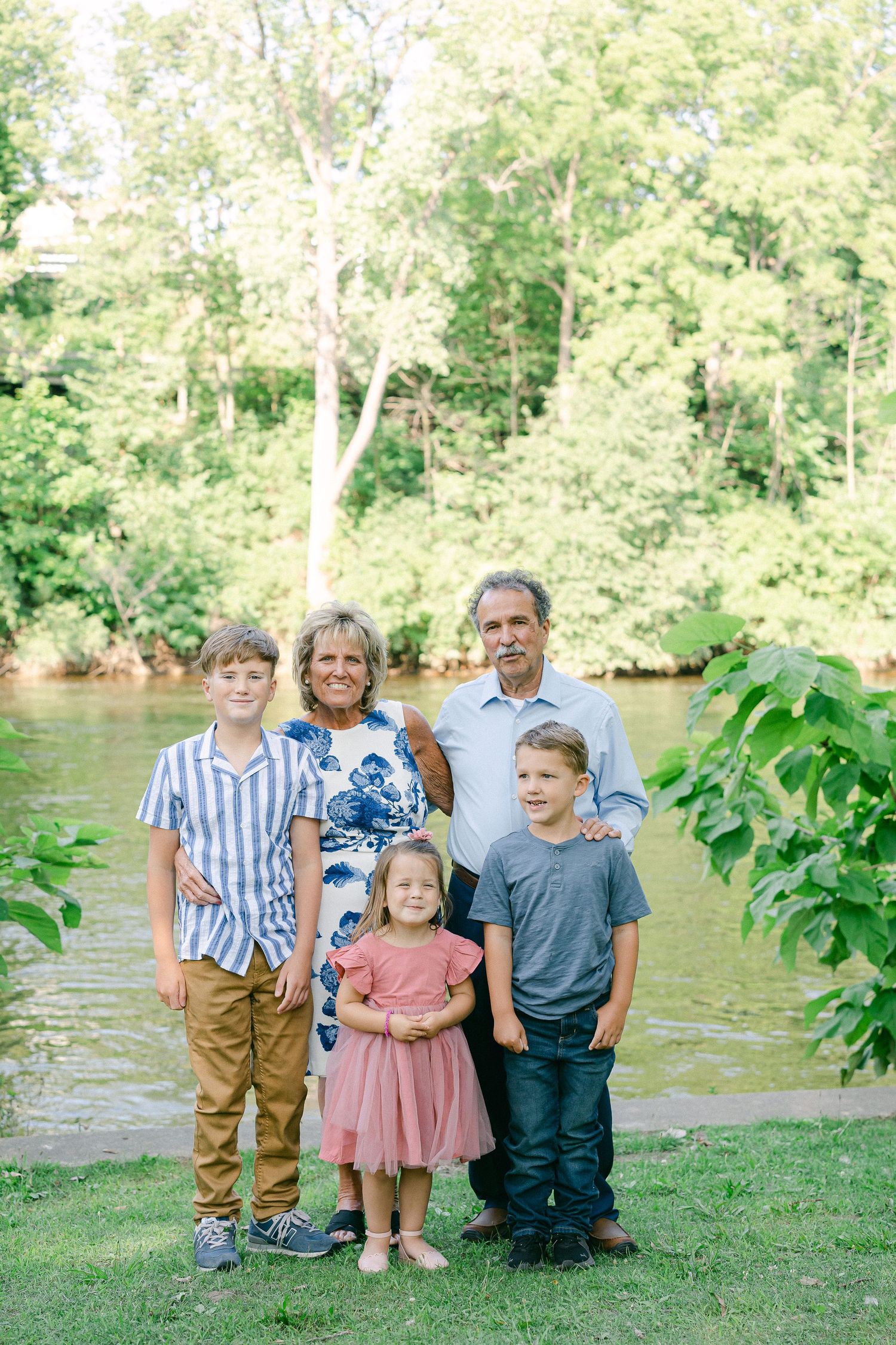 Family group poses together on grassy riverbank during outdoor photo session in natural setting.