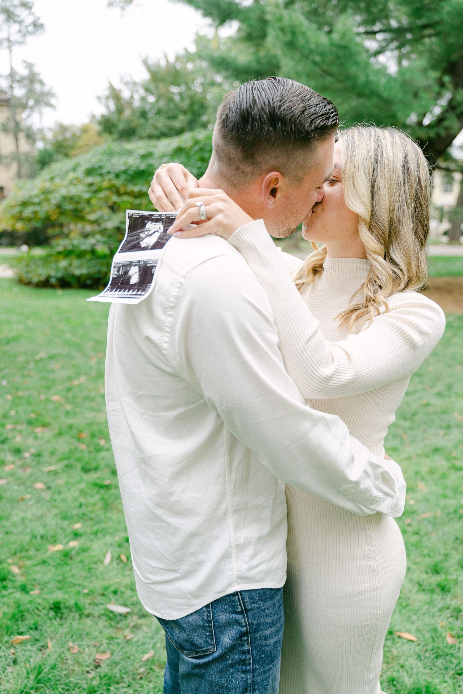 A couple embraces while holding a sonogram photo in a park setting during an outdoor pregnancy announcement photoshoot.