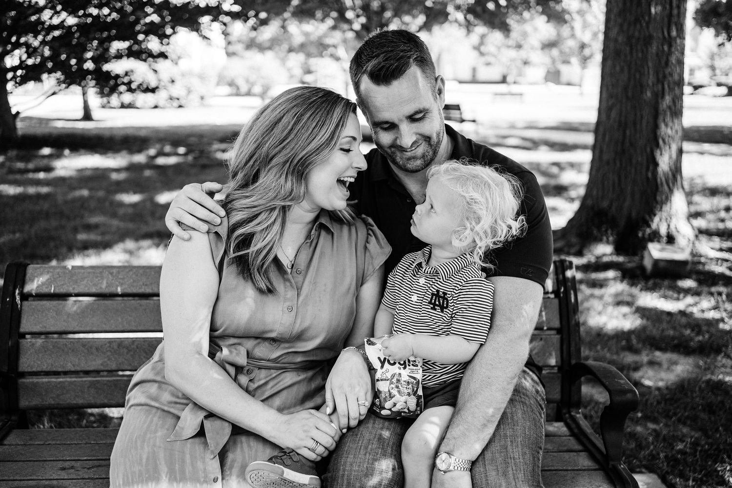 A romantic black and white photo series of a couple sitting on a park bench and embracing in dappled sunlight.
