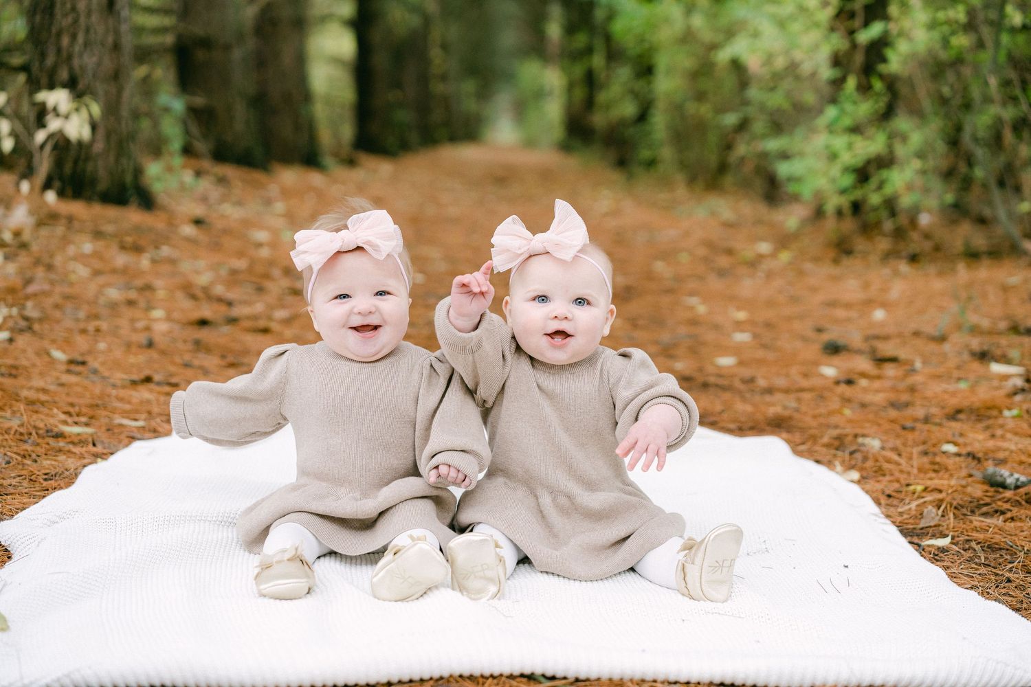 Babies wearing matching beige outfits pose on white blankets in an outdoor autumn setting with fallen leaves.