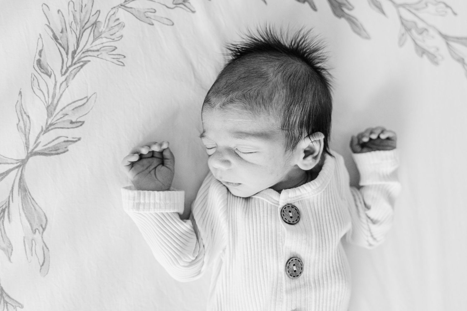 Monochromatic photo of baby lying on white floral background with arms outstretched.