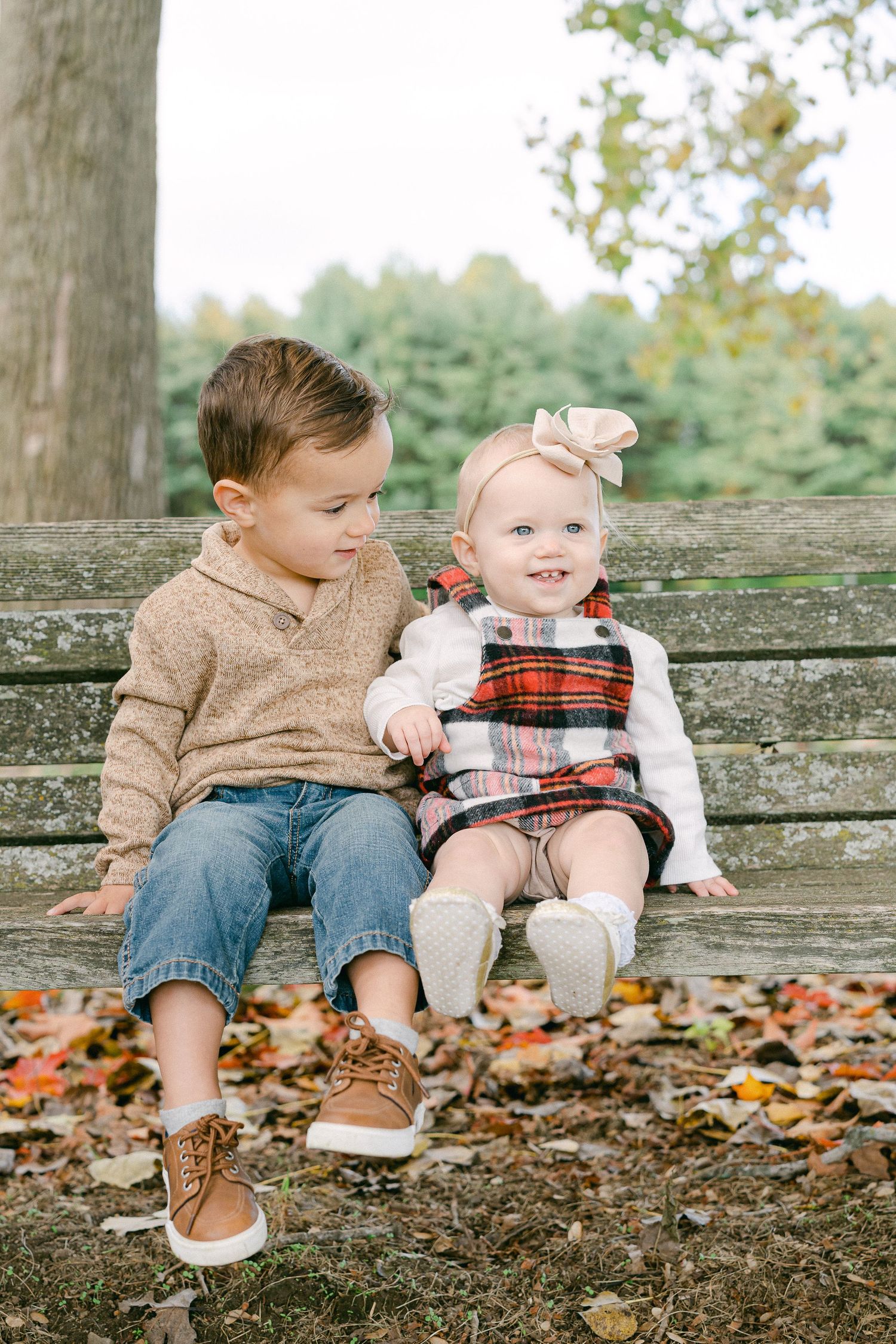 Two young children sit together on a rustic wooden fence surrounded by autumn leaves in an outdoor setting.