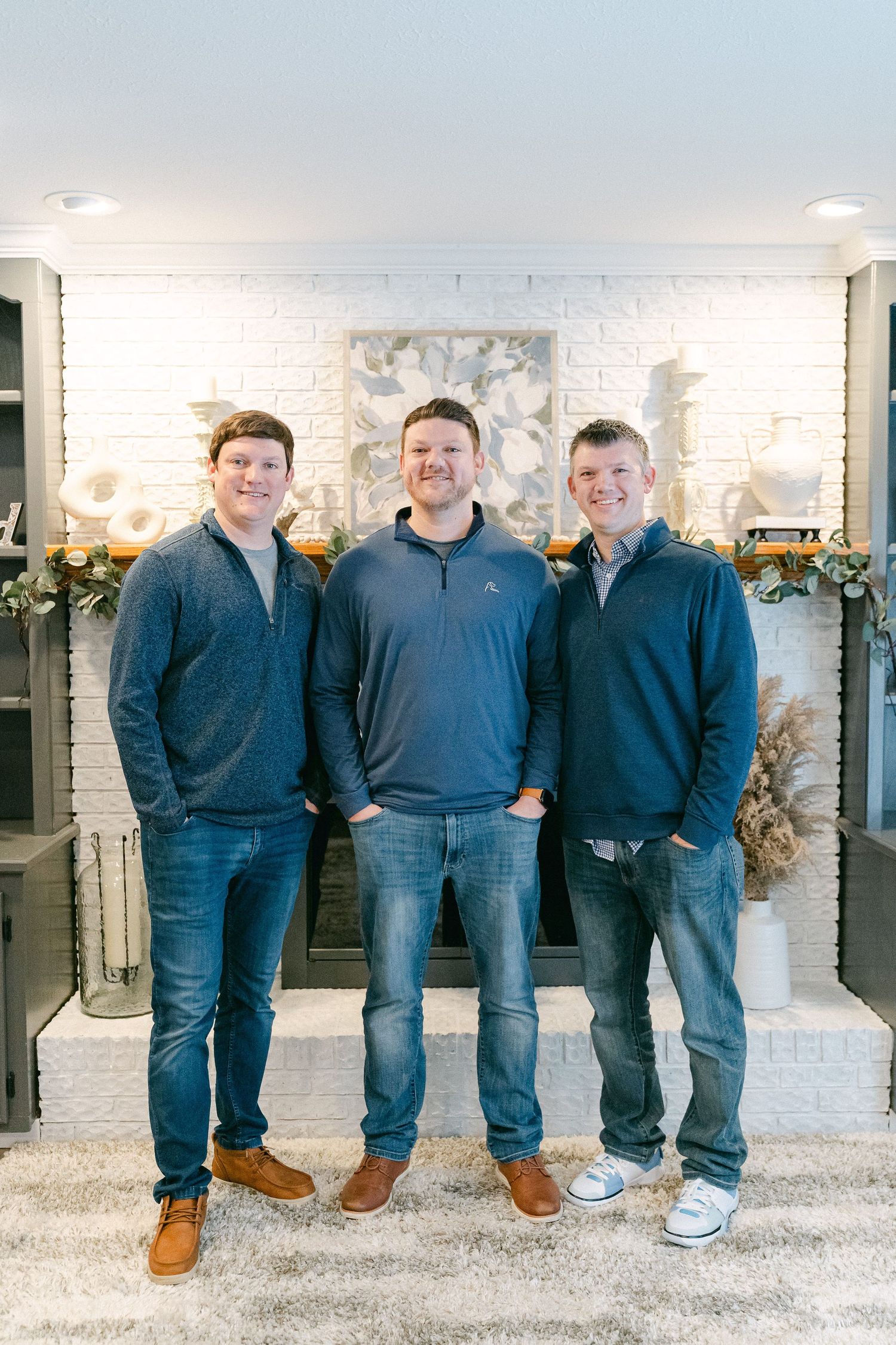 Group wearing matching navy blue outfits and brown shoes stand casually against white brick wall with greenery decor.