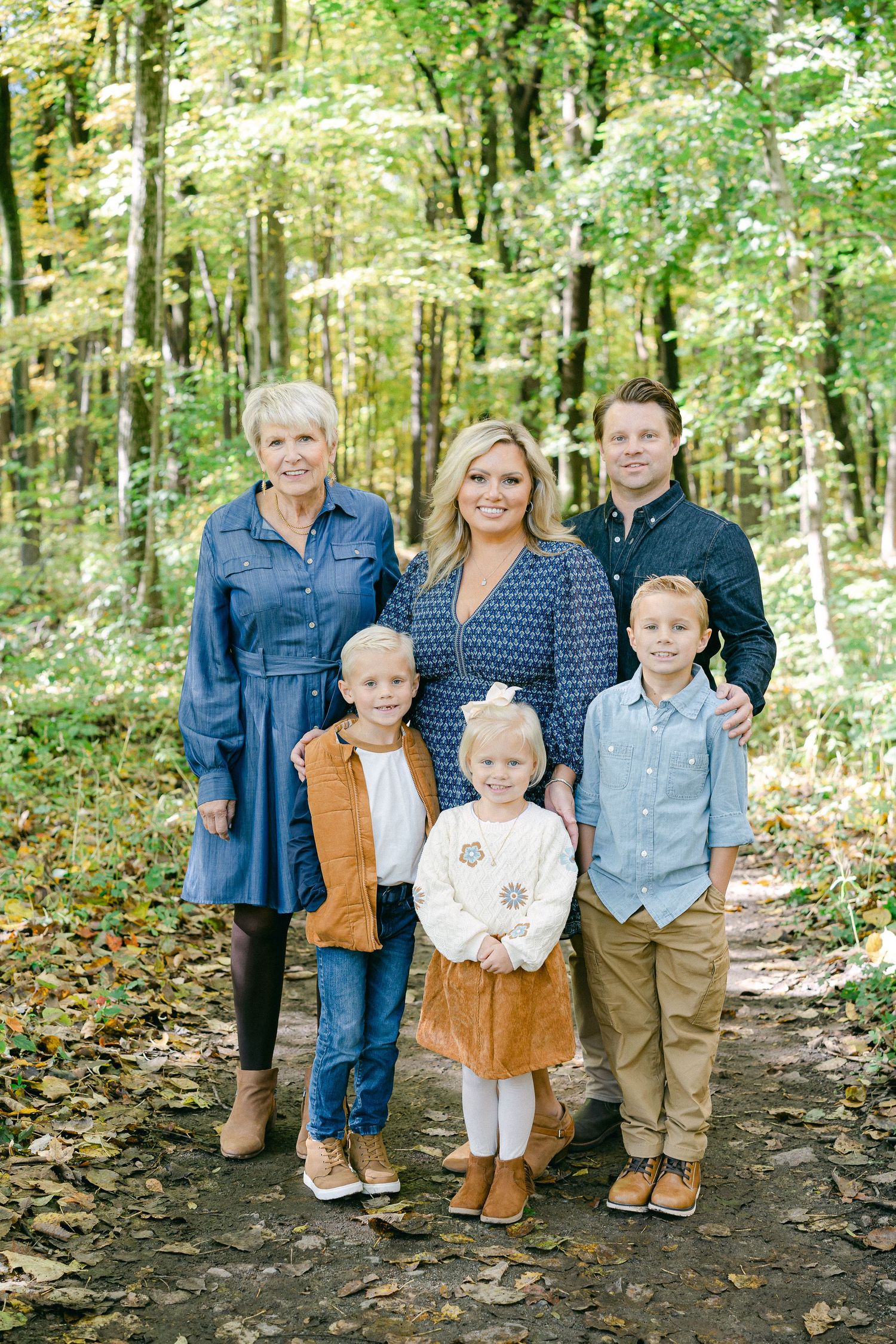 A group poses together on a leaf-covered forest path during autumn wearing coordinated casual outfits in earth tones.