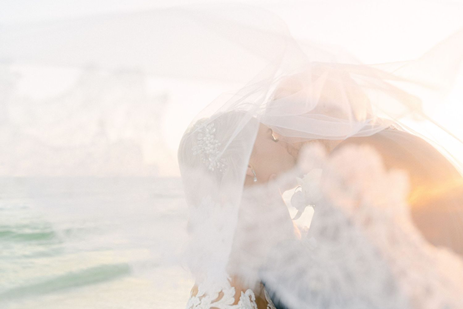 Bride and groom share a romantic kiss on the beach, wrapped in a flowing white veil at golden sunset in Destin, FL