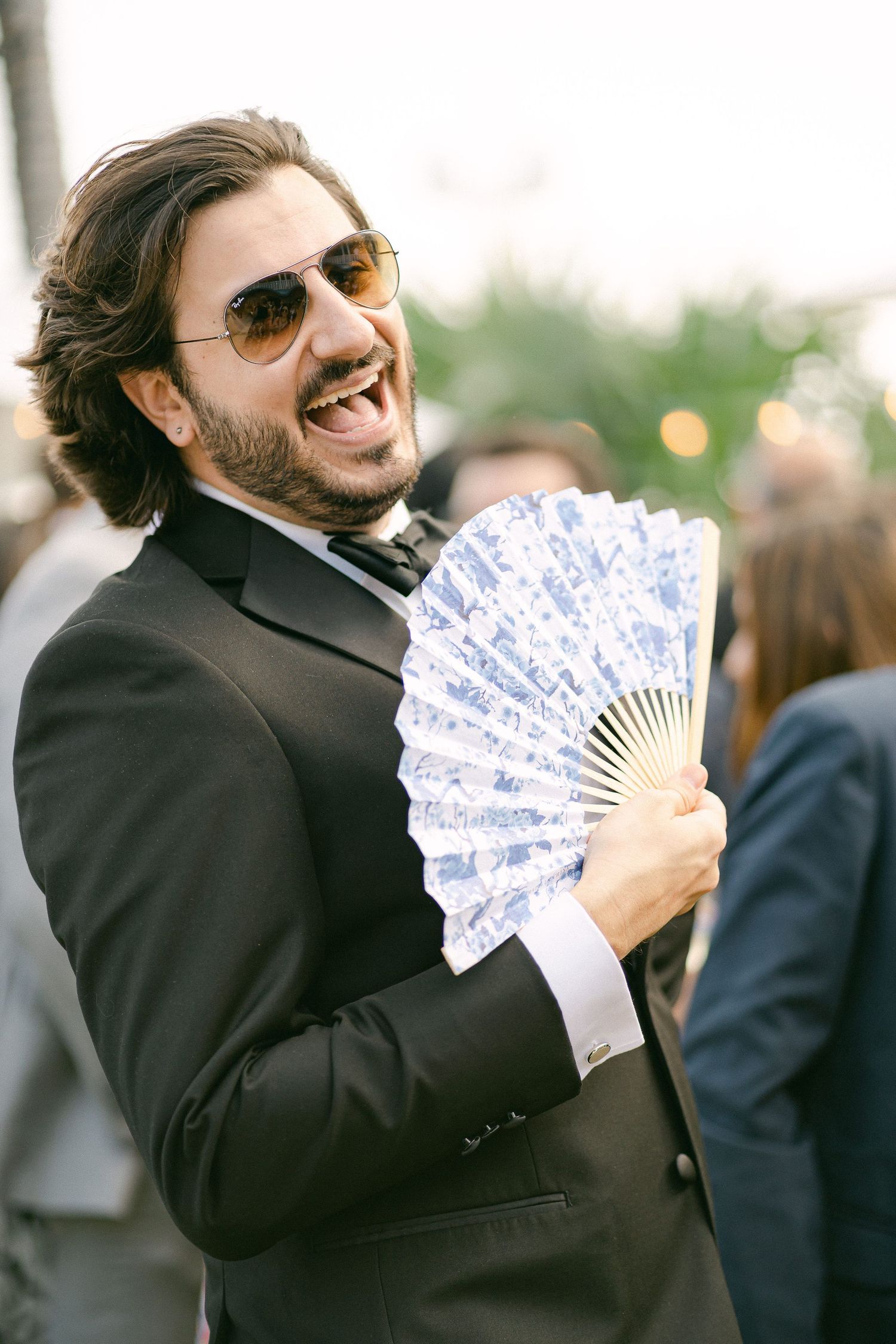 A cheerful bearded man in a black tuxedo and sunglasses holds a decorative blue hand fan at an outdoor event in Miami, FL