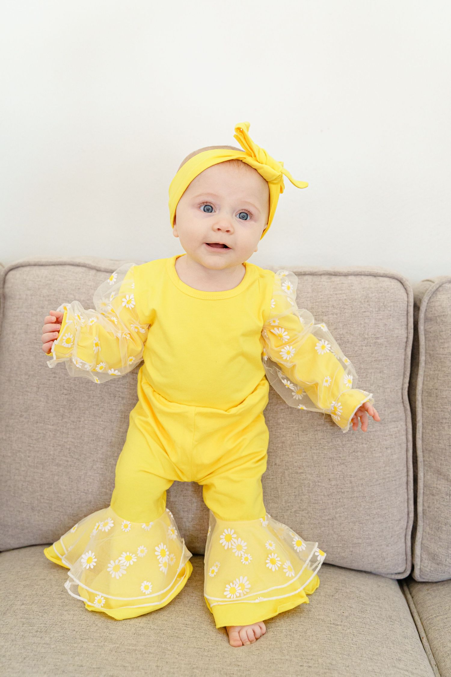 A baby wearing a bright yellow ruffled outfit with white trim sits on a grey couch.